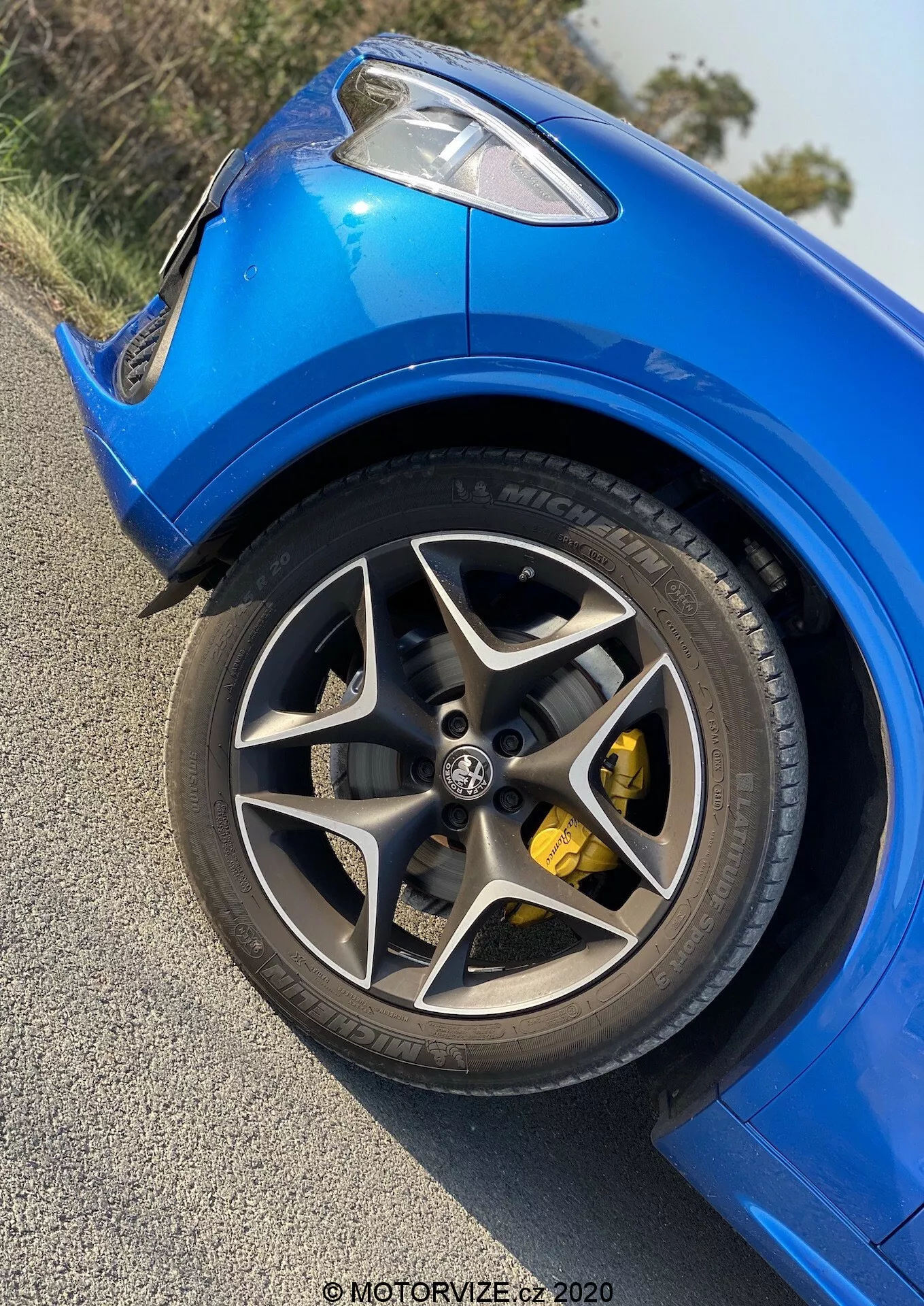 Vista ravvicinata della ruota anteriore sinistra e del parafango dell'Alfa Romeo Stelvio 2017 da un angolo basso, con un focus sullo cerchio in lega multi-razza con finitura scura, un grande disco del freno e una pinza del freno gialla brillante. Si evidenziano le linee dinamiche e i contorni della carrozzeria, la vernice blu brillante e gli pneumatici ad alte prestazioni a profilo basso con scritte bianche. Foto diurna che mostra un'Alfa Romeo Stelvio sportiva e ben mantenuta in condizioni meteorologiche serene; filigrana che legge 'MOTORWIZE.cz 2020'.