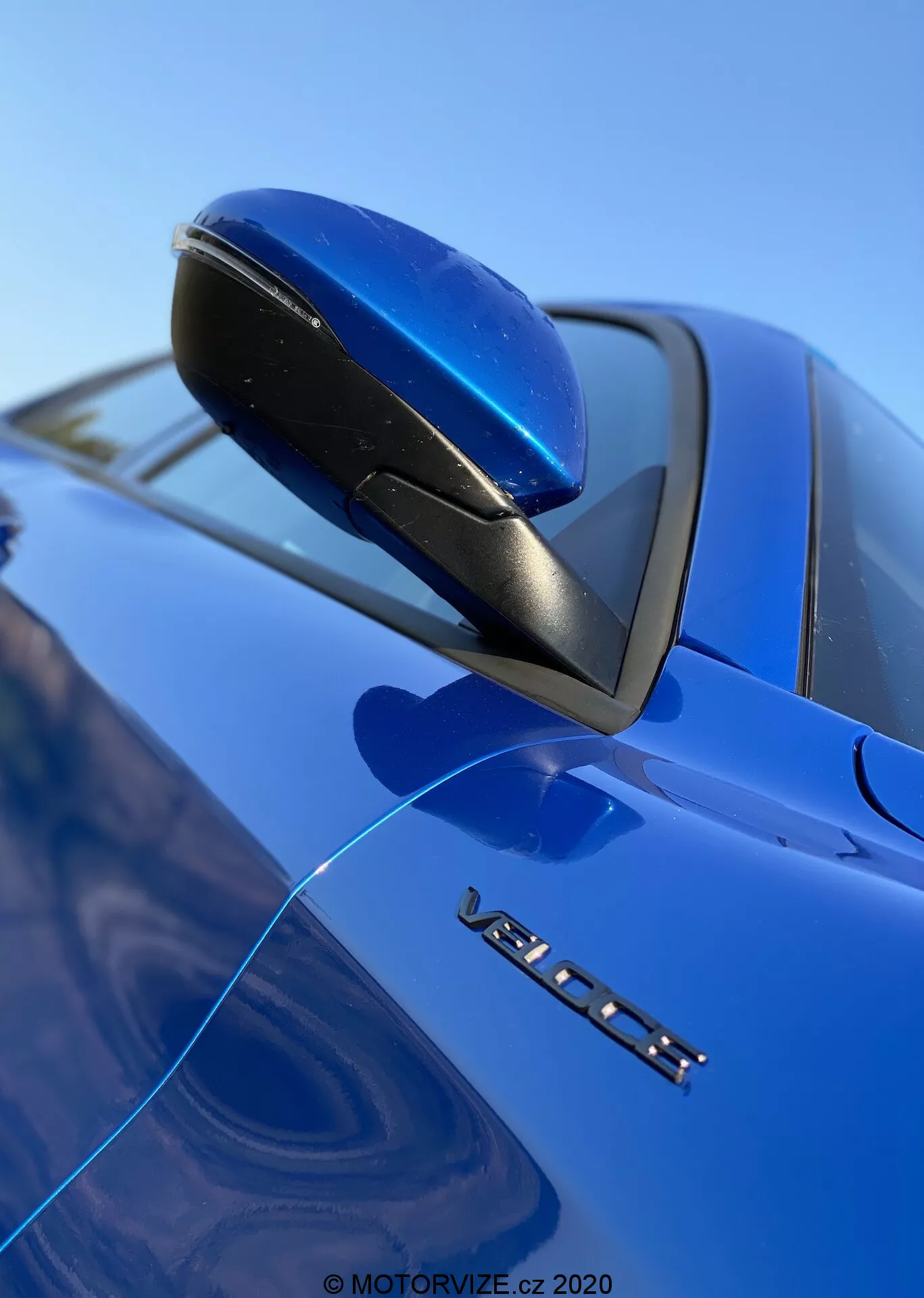 Vista ravvicinata dal basso dello specchietto laterale di una Alfa Romeo Stelvio 2017 blu, che enfatizza il suo design aerodinamico, la base nera e l'indicatore di segnale di svolta integrato. Il marchio VOLVO e il distintivo del modello parzialmente visibile sono visibili sotto lo specchietto, con una messa a fuoco nitida su questi dettagli contro uno sfondo sfocato di parabrezza e cielo, evidenziando la finitura della vernice lucida dell'auto.