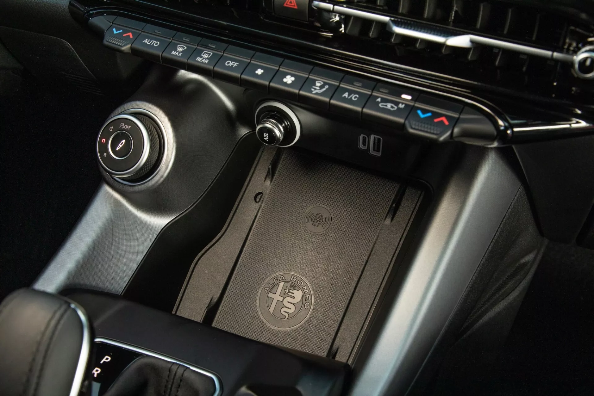 Vista ravvicinata della consolle centrale dell'Alfa Romeo Tonale 2022, che mostra il pannello di controllo del clima, il pad di ricarica wireless, la leva del cambio e il quadrante di controllo dell'infotainment con finitura premium e design moderno.