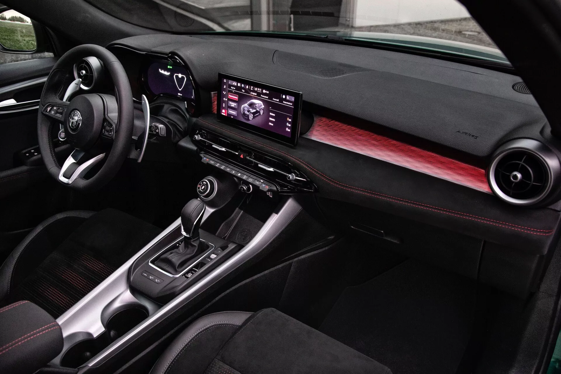 Interno dell'Alfa Romeo Tonale 2022 con cruscotto moderno, centro infotainment digitale, dettagli in fibra di carbonio e sedili con cuciture rosse, visuale dal lato passeggero anteriore