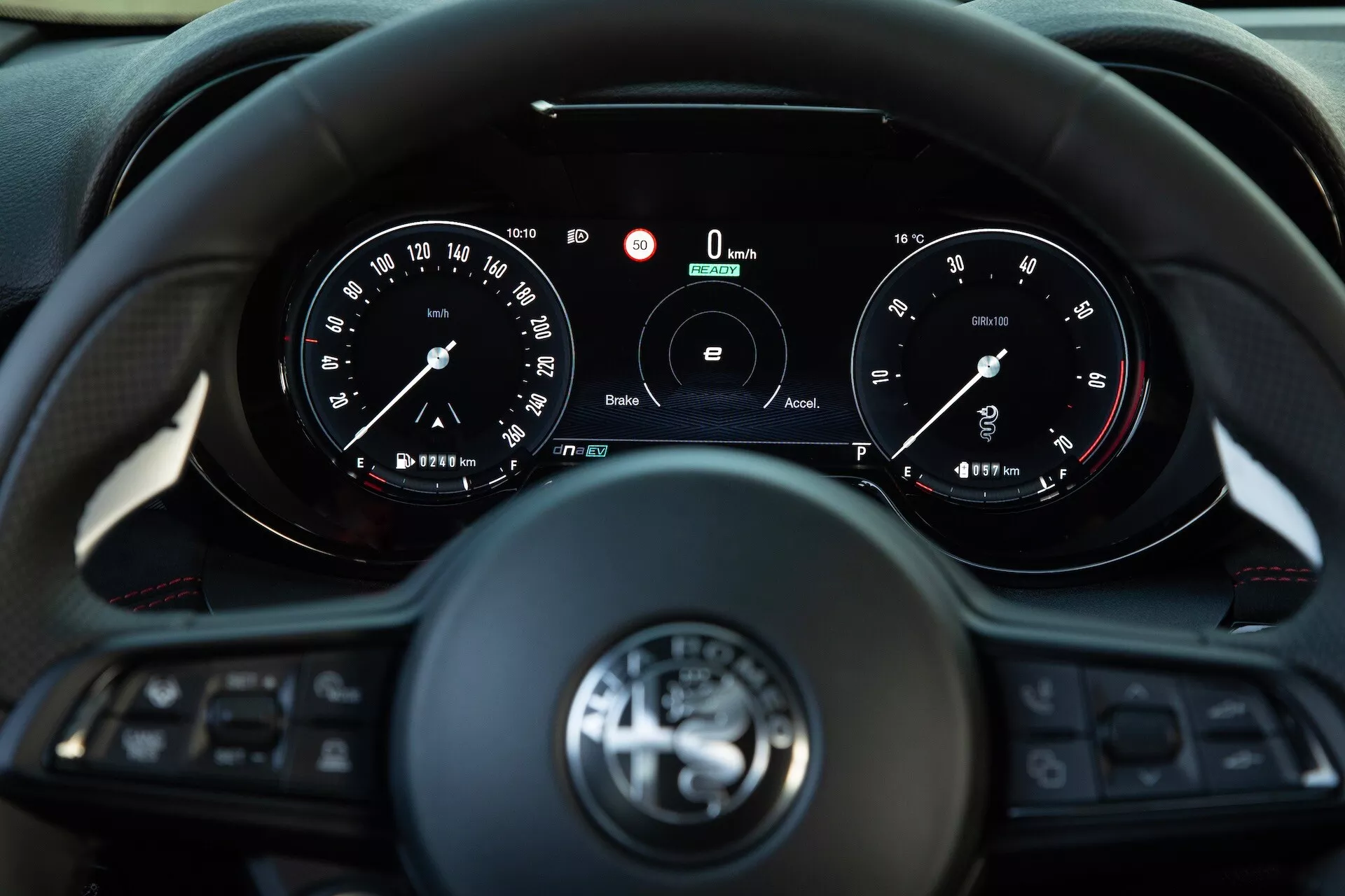 Interni dell'Alfa Romeo Tonale 2022 che mostrano la vista del conducente su volante multifunzione a tre razze e quadro strumenti con indicatori analogici e display digitale centrale che indica velocità e RPM zero, con tecnologie di connettività e assistenza alla guida