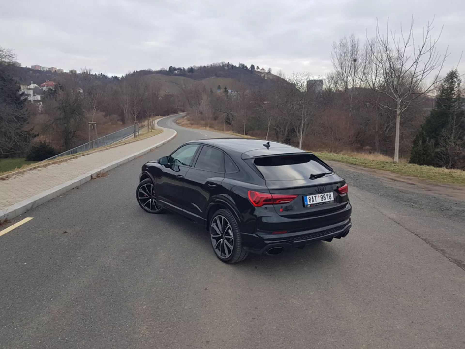 Audi RS-Q3 del 2015 in nero, catturata da una prospettiva posteriore di tre quarti che mette in mostra il suo design sportivo, fanali posteriori LED con luce antinebbia posteriore attivata, doppio scarico, ruote in lega a più razze e spoiler posteriore su uno sfondo suburbano sereno