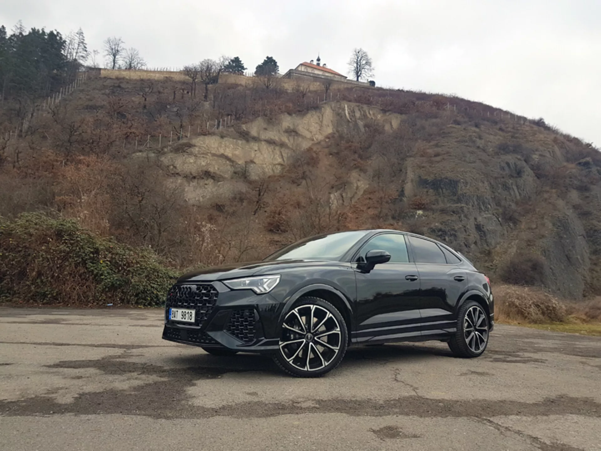 Vista frontale laterale del Audi RS-Q3 2015 parcheggiata all'aperto su un piazzale pavimentato con linee sbiadite, mostrando ruote in lega e griglia anteriore contro uno sfondo collinare nuvoloso