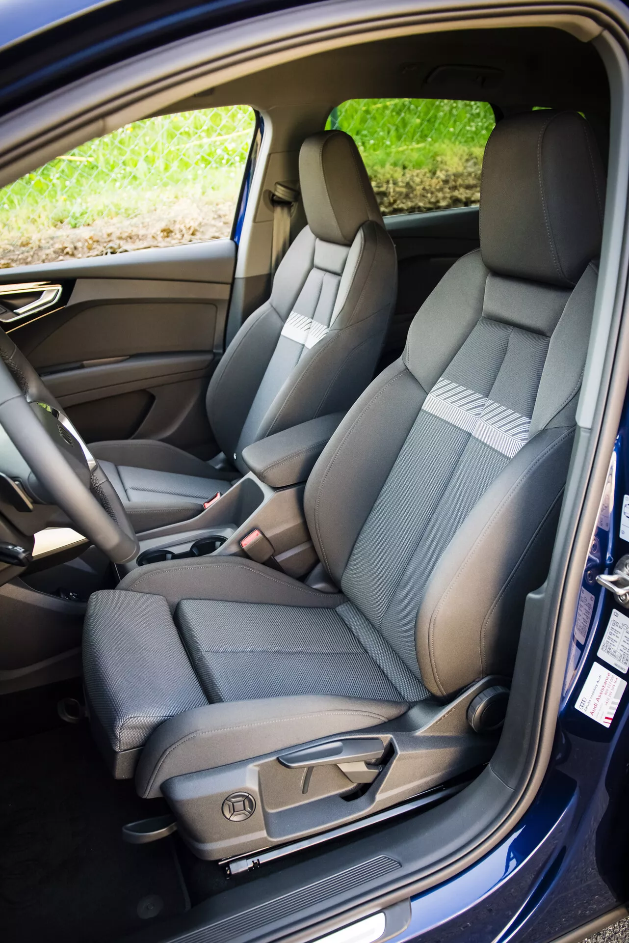 Interni Audi Q4 e-tron 2021 che mostrano il sedile anteriore lato passeggero con tappezzeria bicolore, cuciture dettagliate, comandi per la regolazione elettrica e cintura di sicurezza, accanto a uno scorcio del sedile posteriore con funzione di ribaltamento frazionato e funzioni tecnologiche dell'abitacolo.