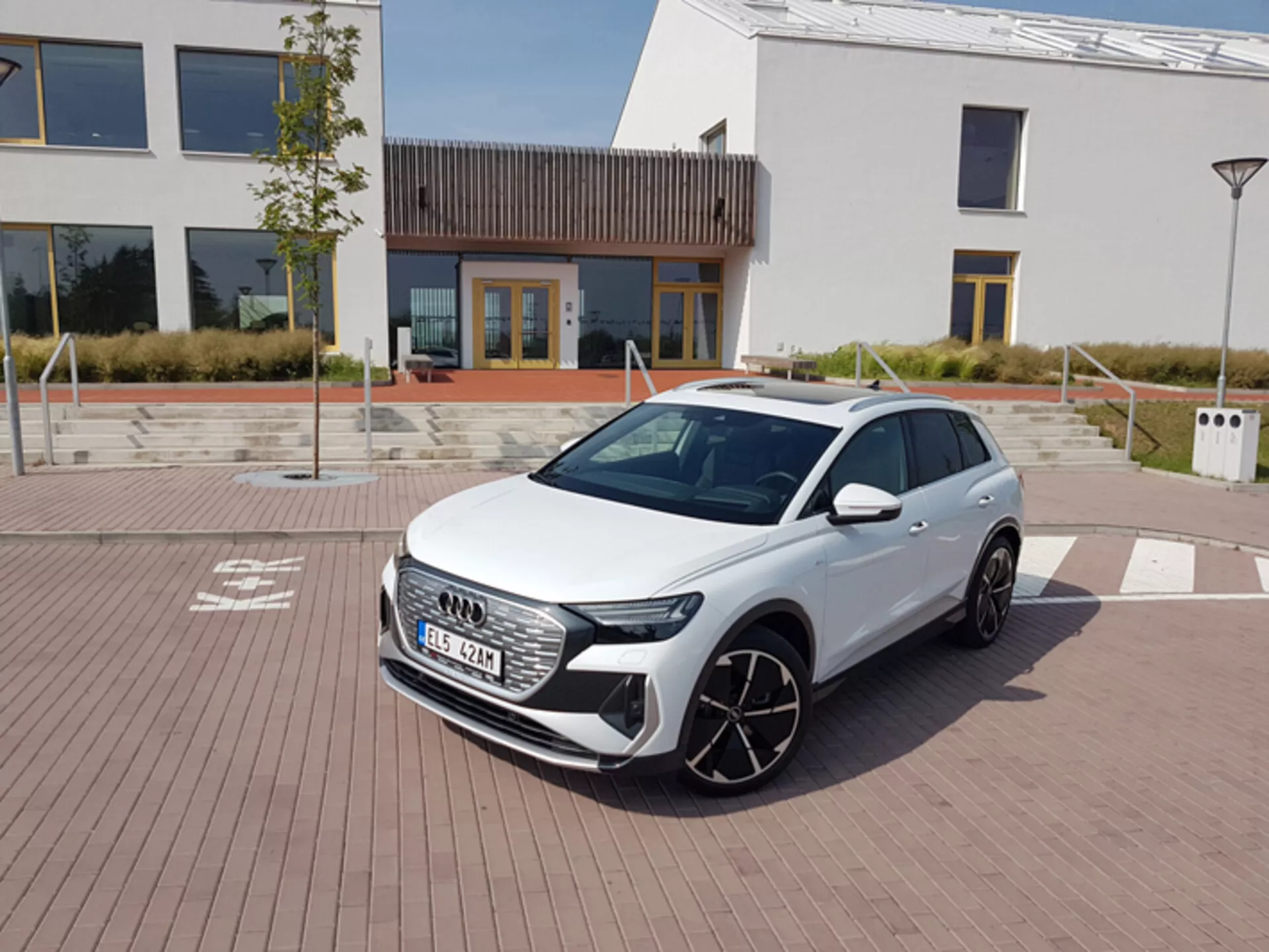 Audi Q4 e-tron 2021 di colore bianco, mostra i suoi moderni fari LED, griglia frontale esagonale e grandi ruote in lega, parcheggiata su un pavimento con motivo a mattoni con architettura urbana e una stazione di ricarica sullo sfondo, sotto un cielo limpido a mezzogiorno.