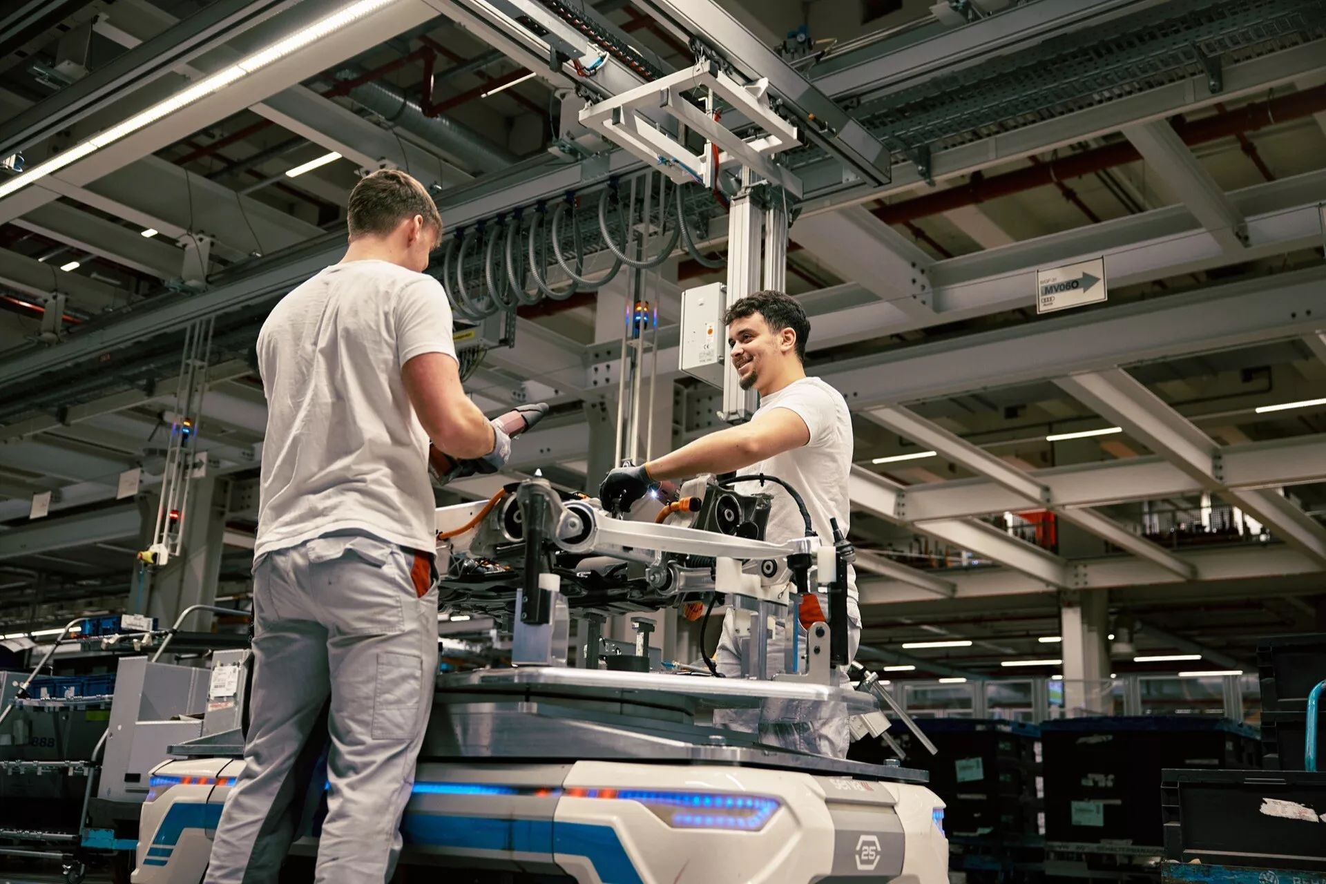 Ambiente industriale con due lavoratori che assemblano o mantengono attrezzature meccaniche sul pavimento di una fabbrica, sotto illuminazione artificiale, con un punto di vista elevato che sottolinea l'ambiente di produzione