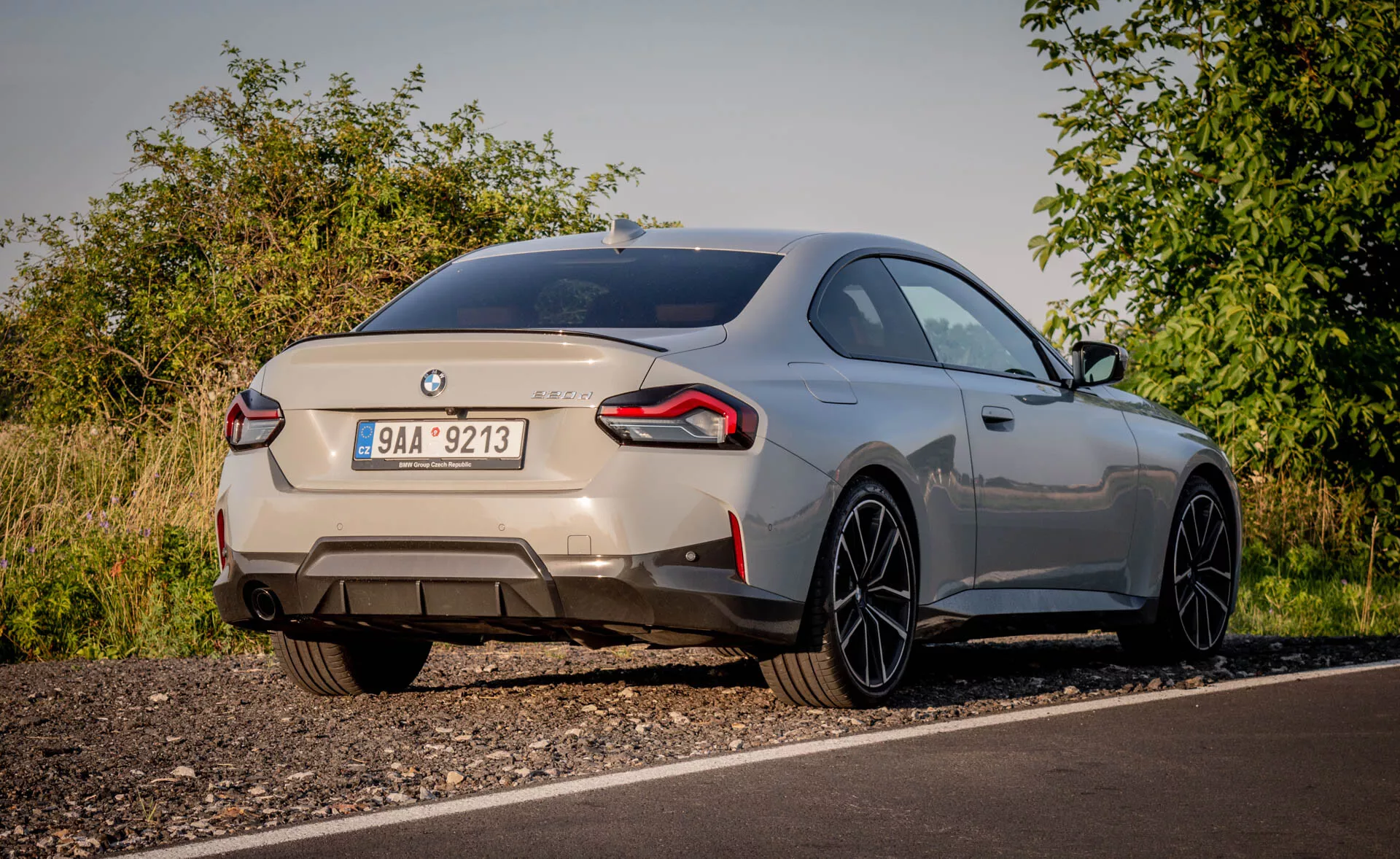 Vista posteriore di una BMW 2er-reihe 2022 (coupé serie 2) sportiva, catturata da una prospettiva angolata bassa. L'auto, parcheggiata sul bordo di una strada in un ambiente rurale con vegetazione naturale, è vista con luce soffusa. I distintivi badge identificano la marca e il modello, mostrando la parte posteriore che include il cofano, il paraurti posteriore e le luci posteriori avvolgenti. I doppi tubi di scarico suggeriscono un motore potente. Un piccolo spoiler posteriore integrato e grandi cerchi in lega con raggi sottili indicano uno stile aerodinamico e performante. La vernice metallica dell'auto e la linea del tetto che scende dolcemente migliorano il suo aspetto premium. Le possibili caratteristiche moderne visibili includono luci posteriori a LED, sensori di parcheggio e una telecamera posteriore.