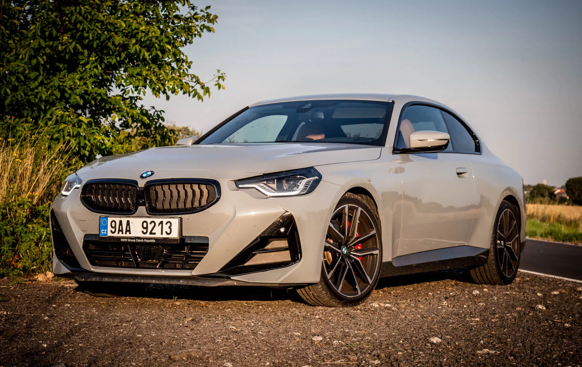 Immagine di una coupé sportiva moderna BMW Serie 2 2022. L'auto è parcheggiata sul ciglio di una strada durante il giorno, vista con un angolo obliquo dalla parte anteriore sinistra, evidenziando il lato del guidatore. Le caratteristiche includono un design aerodinamico elegante, grandi ruote in lega multirazza con finitura scura, pinze dei freni in contrasto, una griglia distintiva affiancata da fari LED stretti e aggressivi, una telecamera frontale sopra la targa, grandi prese d'aria nel paraurti anteriore, vernice metallica chiara, specchietti laterali con indicatori di svolta integrati e una vista parziale del tetto panoramico. L'auto è posizionata vicino alla fotocamera con uno sfondo rurale o suburbano, illuminazione calda e ombre che indicano un sole basso al mattino o nel tardo pomeriggio.