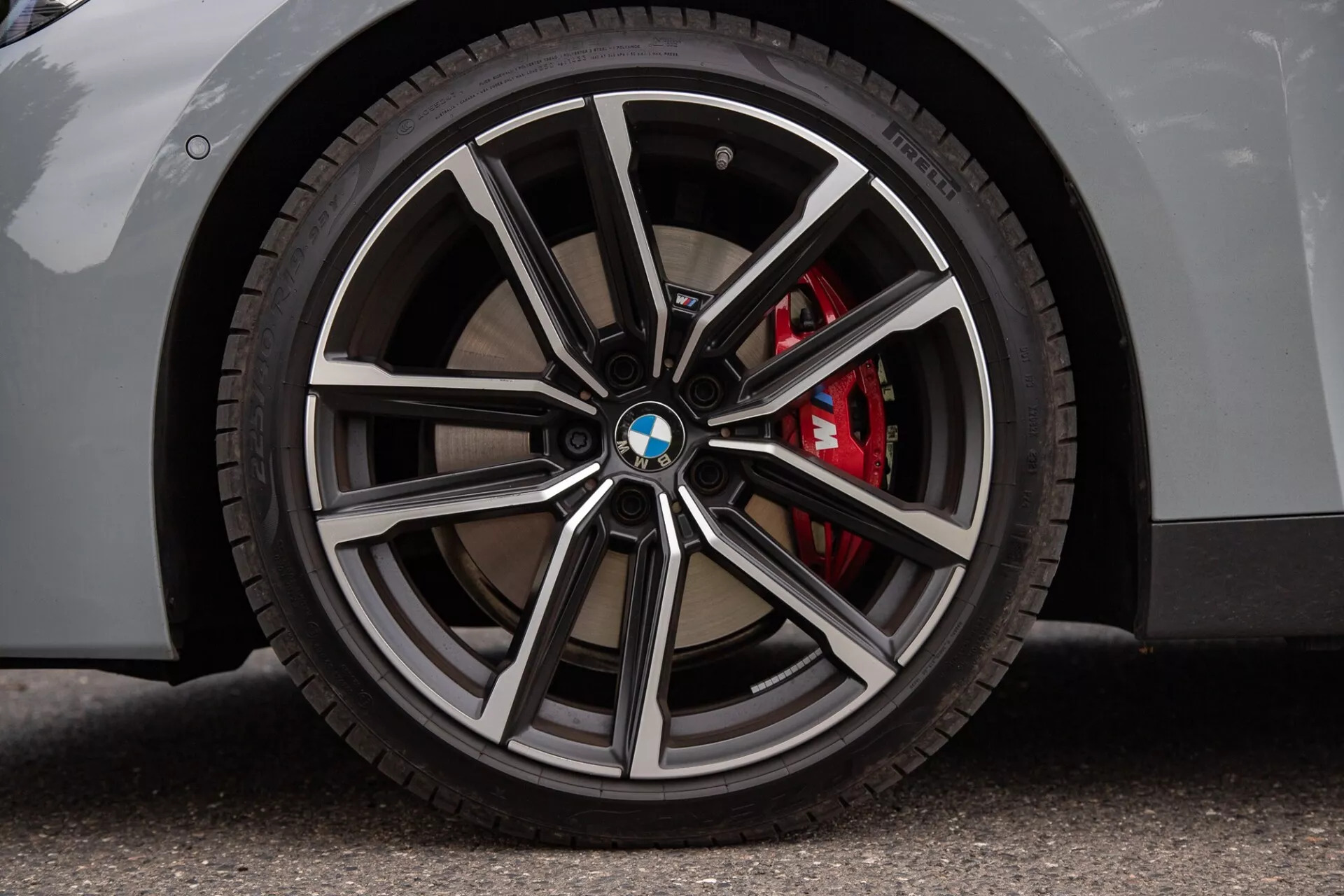 Primo piano della ruota di una BMW Serie 2 Coupé che mostra un cerchio multirazza con finiture metalliche lucide e scure, logo BMW sul coprimozzo, pneumatico a profilo basso e pinza dei freni rossa ad alte prestazioni. La carrozzeria chiara su superficie asfaltata enfatizza gli aspetti di lusso e sportività.