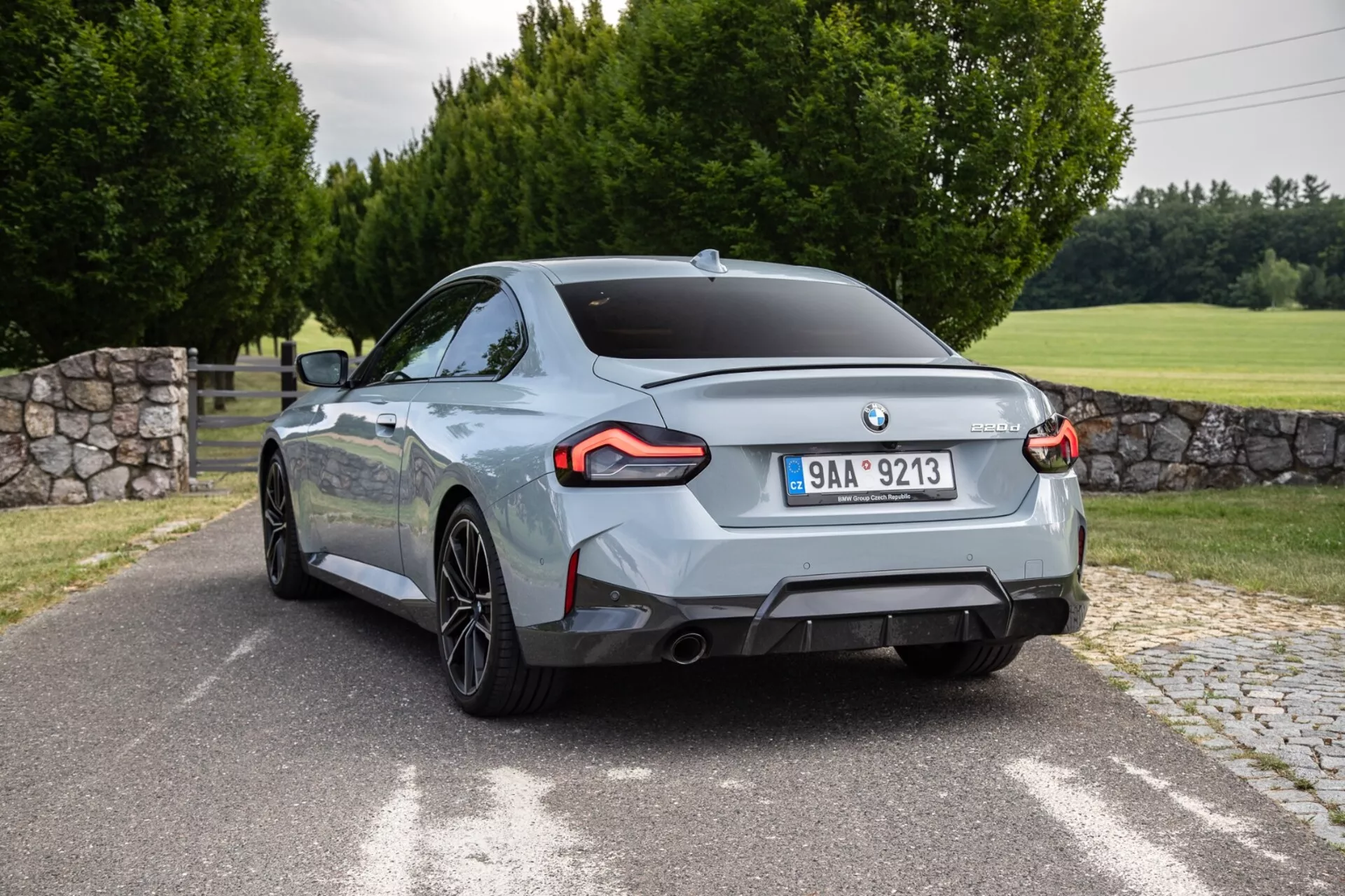 Immagine della BMW 2er-Reihe 2022 2-Series Coupe catturata da un'angolazione posteriore di tre quarti. L'auto, posizionata quasi diagonalmente, mostra caratteristici fanali posteriori a LED angolari, doppi terminali di scarico, cerchi in lega a più raggi e una finitura di vernice metallica lucida. Il design posteriore presenta un emblema del produttore centrato e un distintivo della versione del modello/motore sul cofano del bagagliaio. L'ambiente esterno include una strada pavimentata o vialetto, area erbosa, alberi e un muro di pietra parzialmente visibile.
