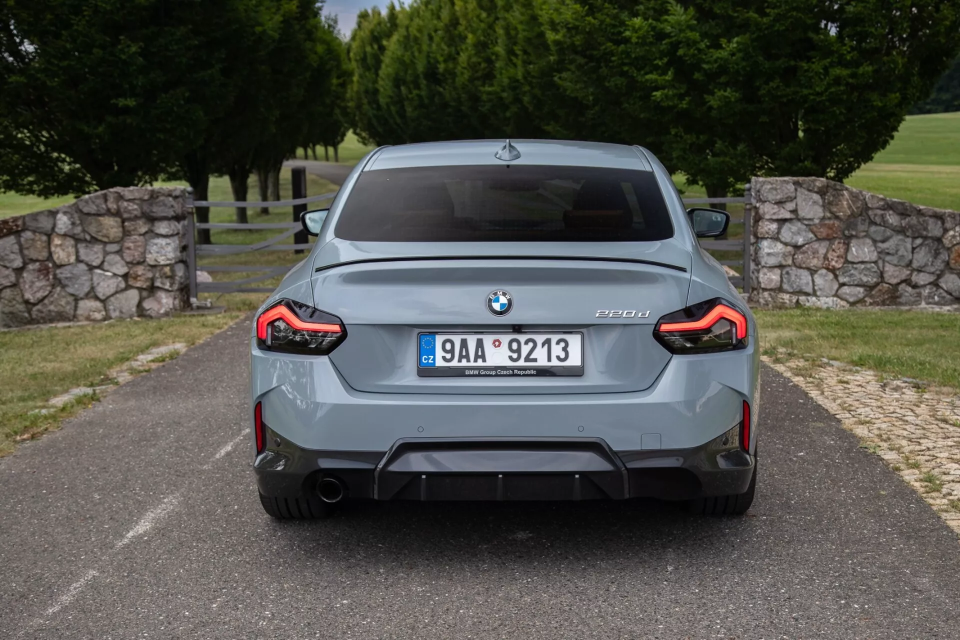 Vista posteriore di una BMW M3 berlina grigia (modello G80) che mostra l'emblema BMW, la targhetta M3 con le informazioni sul modello G80, targa centrale, paraurti posteriore sportivo con diffusore integrato, doppi terminali di scarico, fanali posteriori a LED a forma di L a tutta larghezza e archi passaruota svasati.
