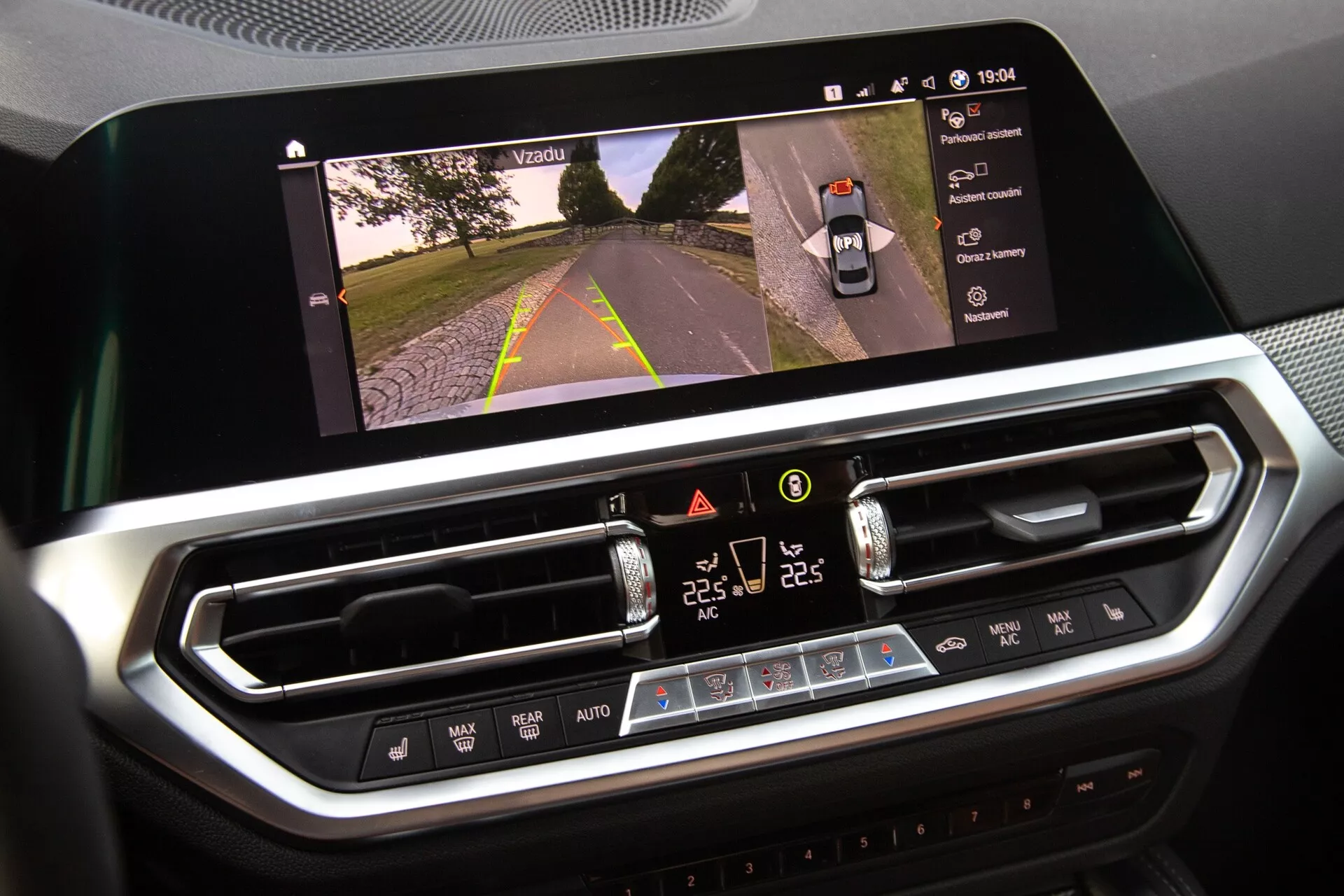 Primo piano degli interni della BMW 2er-Reihe 2022, che mostra il display dell'infotainment e il sistema di controllo del clima. Il grande display orizzontale mostra una visuale della strada ad indicare l'attivazione della telecamera, mentre l'interfaccia di controllo del clima digitale sottostante presenta impostazioni di temperatura a doppia zona, velocità della ventola e pulsanti fisici. Design moderno con finiture cromate che suggeriscono materiali di pregio.