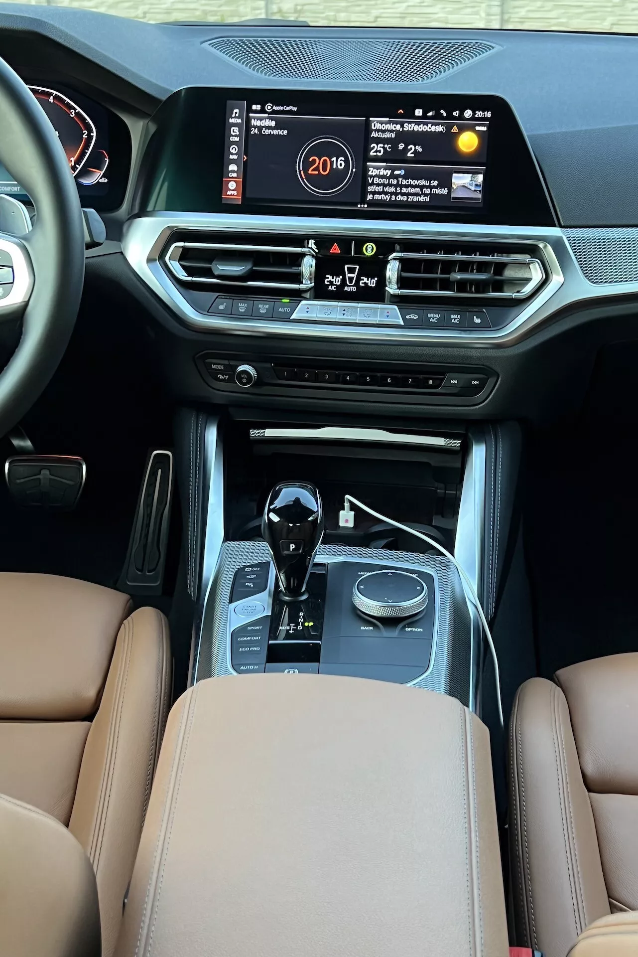 Vista interna della BMW Serie 2 2022 dall'area dei passeggeri posteriori, evidenziando la console centrale con grande schermo di infotainment che mostra ora, temperatura e sistema multimediale. Le caratteristiche includono bocchette di aria condizionata, pulsanti di controllo del clima, pulsante di avvio, leva del cambio automatico con accenti cromati, selettori delle modalità di guida, sedili in pelle marrone con rivestimenti di alta qualità e cuciture, che indicano lusso e tecnologia automobilistica avanzata.