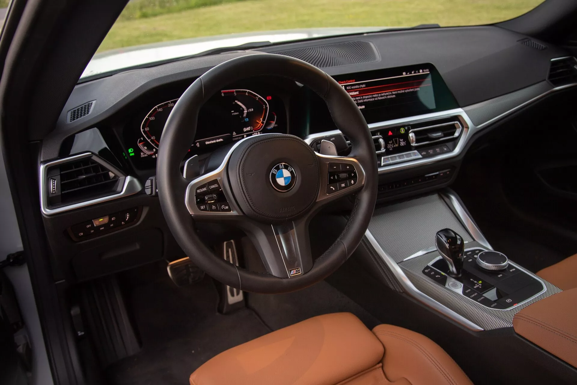 Immagine degli interni di una BMW Serie 2 Coupé 2022 focalizzata sul posto di guida. La ripresa elevata offre una vista dettagliata del volante multifunzionale con logo BMW, quadro strumenti digitale che mostra tachimetro e contagiri, e consol centrale orientata al guidatore con ampio schermo di infotainment, pulsanti di controllo del clima, leva del cambio e comando iDrive. I sedili sono rivestiti in pelle color caramello o marrone chiaro, enfatizzando il lusso e il comfort.