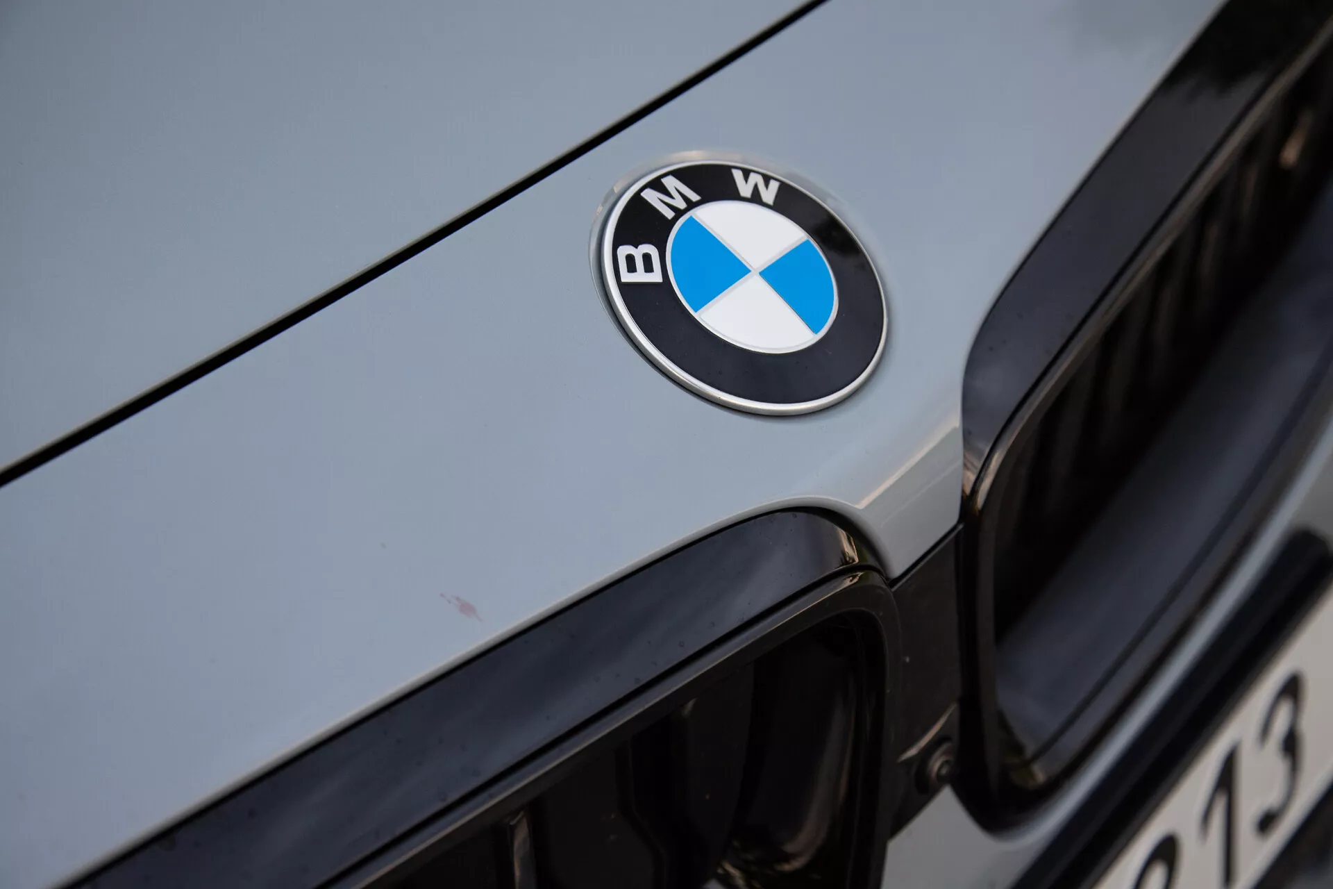 Immagine ravvicinata del frontale della BMW Serie 2 Coupé 2022, con il distintivo BMW nero, blu e bianco ben centrato sul cofano. L'immagine cattura il bordo superiore della griglia a rene iconica sullo sfondo, con un esterno dell'auto di colore argento metallizzato o grigio chiaro che riflette la luce ambientale. La messa a fuoco nitida sul distintivo enfatizza il branding di lusso e la superficie ben mantenuta del veicolo.