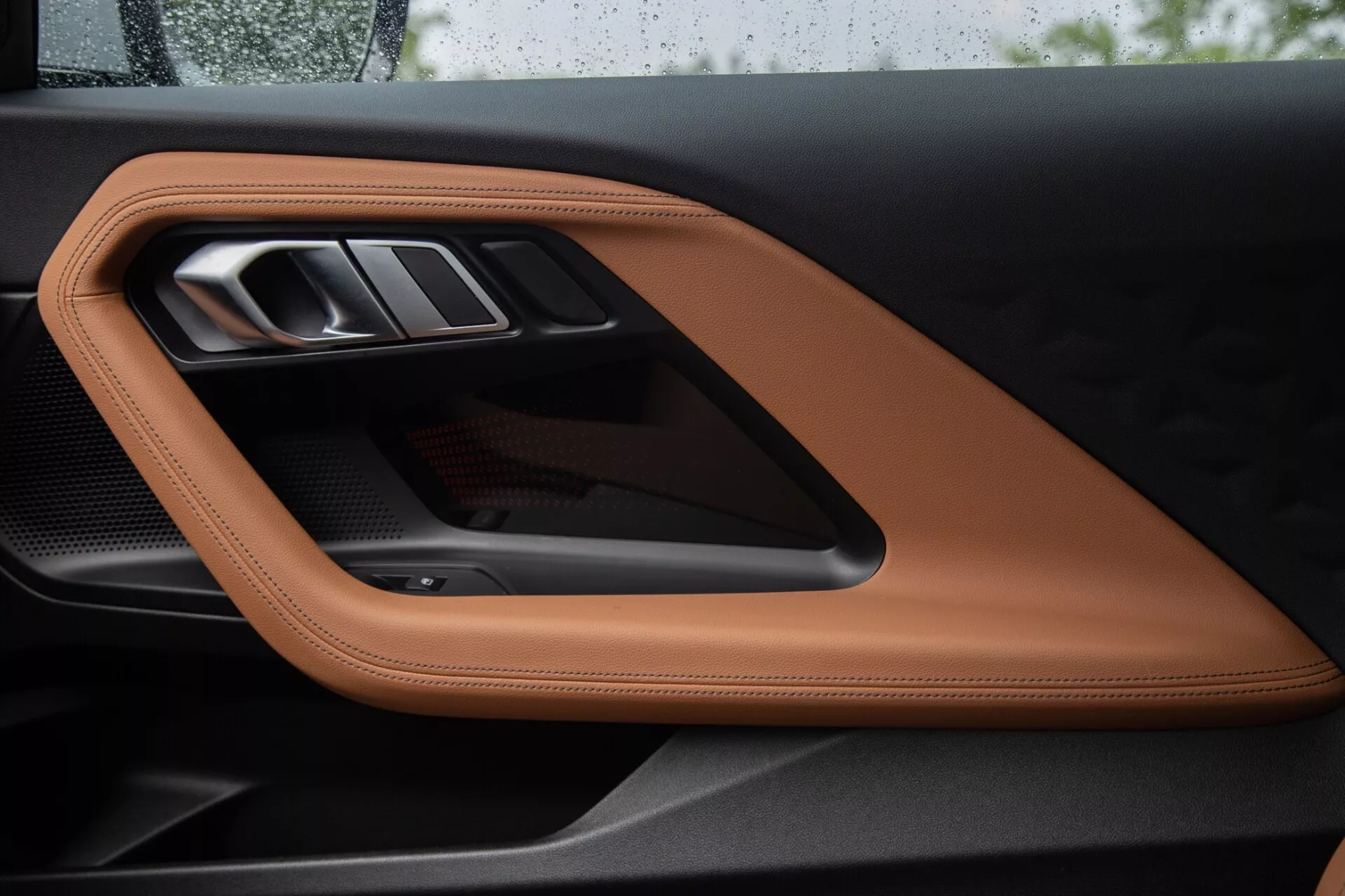 Vista ravvicinata del pannello della porta interna della BMW Serie 2 Coupé 2022, che mostra pelle cucita di lusso color cuoio o cognac, una maniglia della porta metallica elegante e un accento sportivo in fibra di carbonio, evidenziando materiali di alta qualità e elementi di design sofisticati.