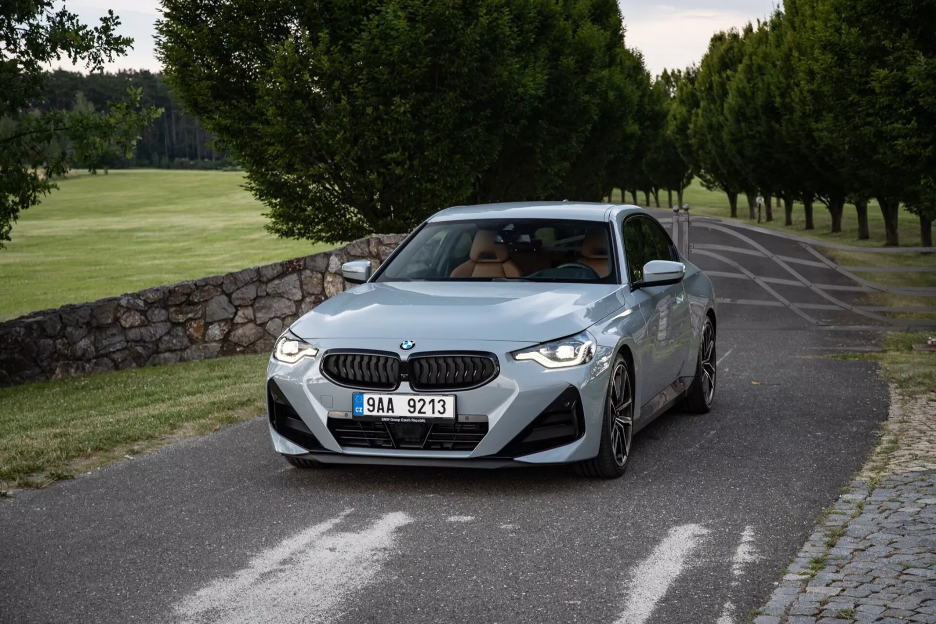 Immagine dettagliata di una BMW Serie 2 Coupé del 2022 catturata da un angolo frontale a tre quarti, che mostra i profili frontale e laterale sulla strada asfaltata serena, fiancheggiata da alberi ben curati e un muro di pietra. La caratteristica griglia a doppio rene BMW e i fari LED eleganti adornano la fascia anteriore, mentre le prese d'aria sul paraurti migliorano l'aspetto sportivo. Le linee sagomate del cofano suggeriscono prestazioni, i parafanghi ben definiti indicano potenza e i cerchi in lega multiraggio denotano un assetto sportivo. Il profilo laterale presenta una linea caratteriale che dà una sensazione di movimento, specchietti retrovisori con indicatori integrati e un accenno di tetto apribile. L'angolo del parabrezza standard e i finestrini laterali chiari aggiungono un aspetto equilibrato. L'illuminazione morbida, probabilmente al mattino presto o nel tardo pomeriggio, crea ombre sottili e una presentazione pulita del design e del colore dell'auto.