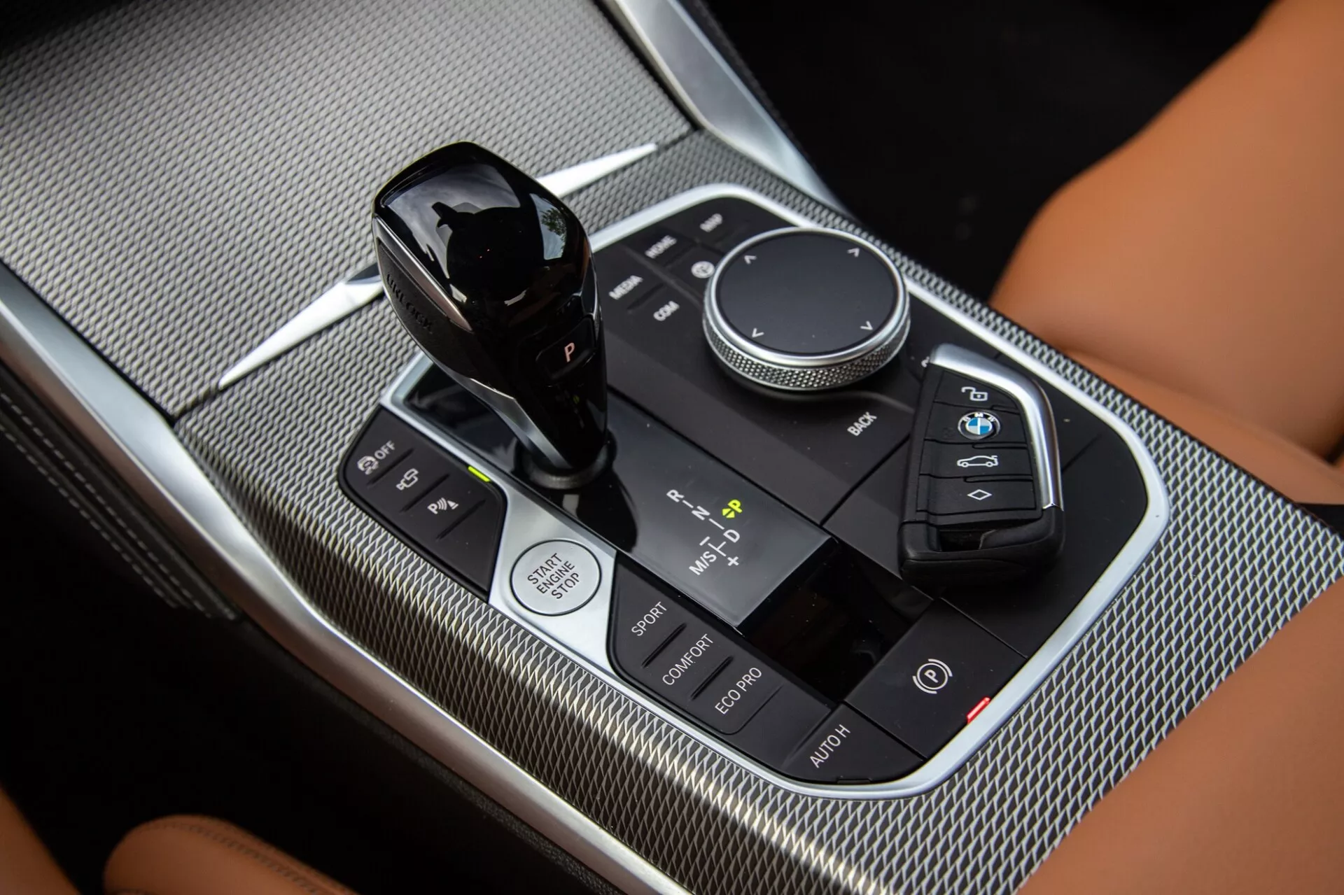 Primo piano della console centrale della BMW Serie 2 Coupé 2022, con evidenza della leva del cambio, della manopola di controllo multimediale e del pannello di controllo touch con finitura nera lucida e superfici con trama in fibra di carbonio.