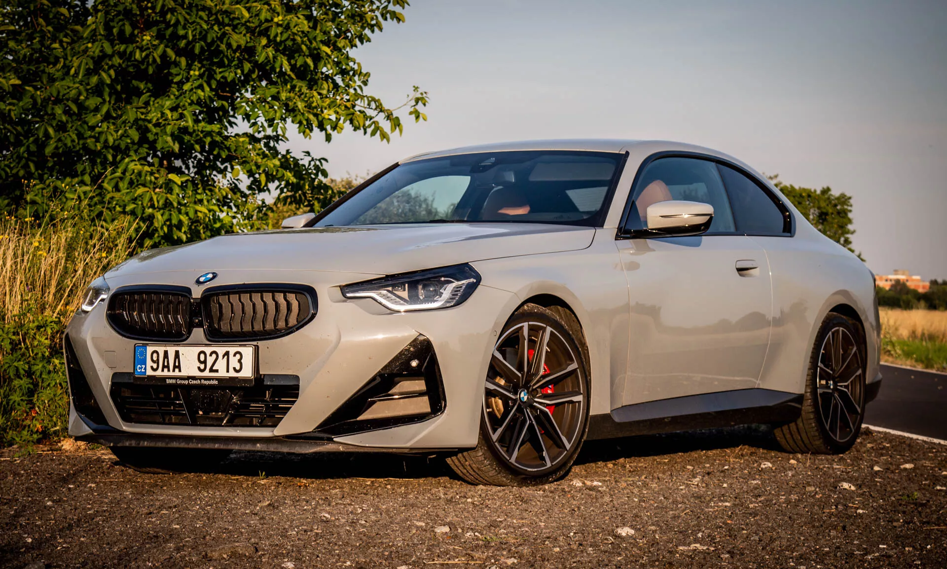 Immagine di una BMW Serie 2 Coupé del 2022. L'auto di lusso moderna è parcheggiata angolata, evidenziando il lato anteriore sinistro durante l'ora dorata. L'inquadratura si concentra su elementi distintivi del design come la grande griglia a forma di rene, i fari LED angolari, il paraurti anteriore sportivo con grandi prese d'aria e i cerchi in lega neri multirazze con pinze dei freni ad alte prestazioni. Lo specchietto laterale aerodinamico ha indicatori di direzione integrati. La carrozzeria elegante e la finitura premium della vernice enfatizzano il profilo compatto, sportivo e l'abbassamento del tetto dell'auto.
