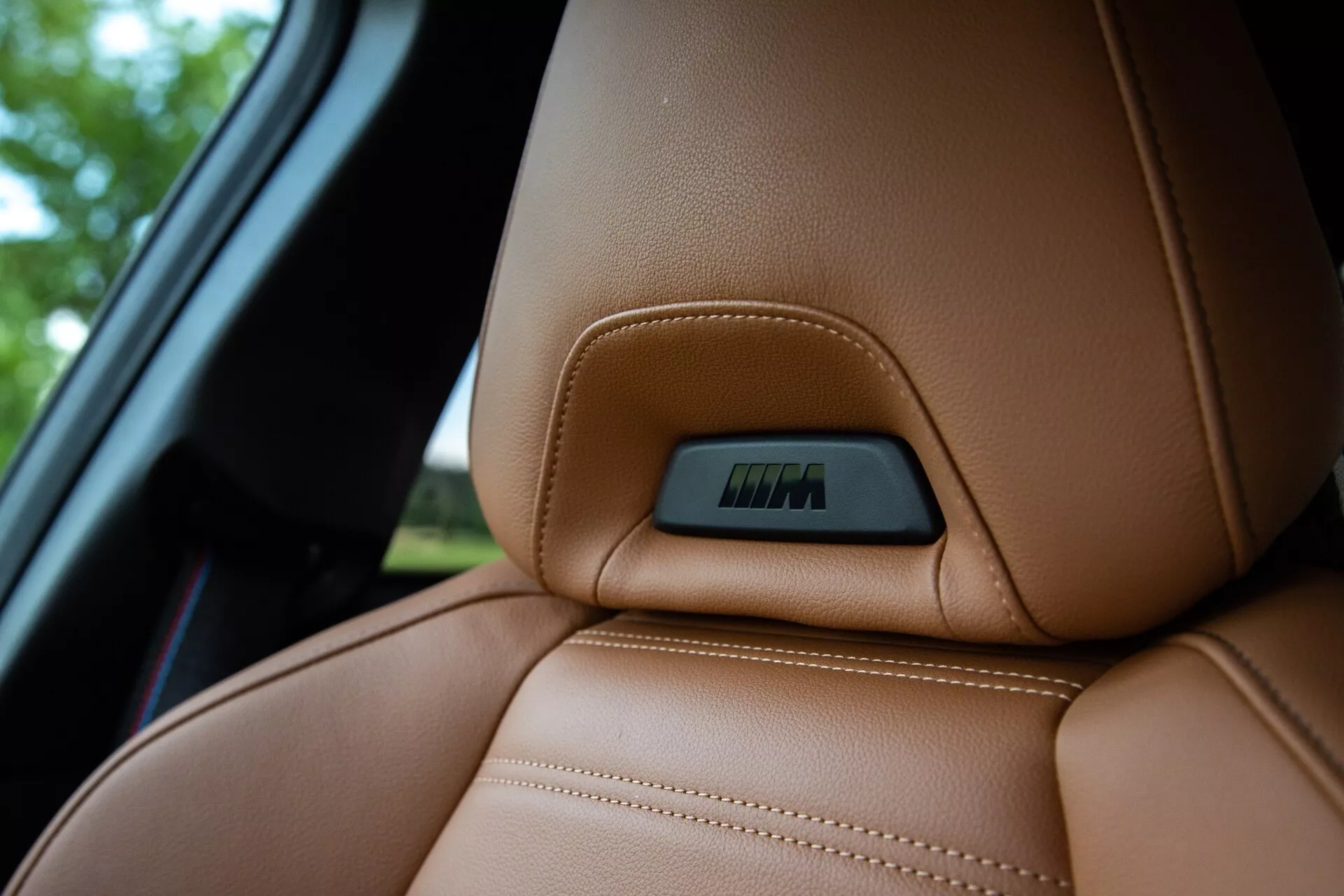 Vista ravvicinata della sezione superiore di un sedile in pelle marrone scuro in una BMW Serie 2 Coupé 2022, evidenziando cuciture visibili precise e una linea di contorno ergonomica per comfort e supporto. Un badge incastonato in una rientranza sulla parte superiore del sedile indica caratteristiche tecnologiche come regolazioni elettroniche o funzioni di riscaldamento. La ripresa è effettuata da una posizione leggermente più bassa, accentuando la texture lussuosa della pelle mentre lo sfondo è sfocato, includendo una porzione del finestrino dell'auto.