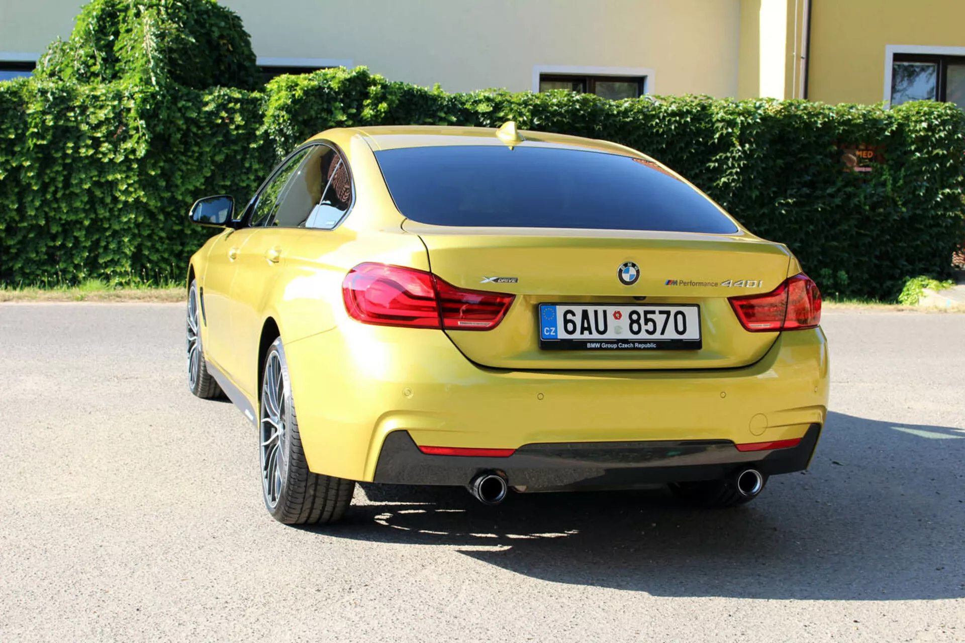 BMW Serie 4 Coupé gialla del 2013 vista da un angolo posteriore di tre quarti che mostra il design del bagagliaio, il paraurti posteriore, parte del passaruota posteriore, luci posteriori distintive, tubi di scarico doppi, targa, emblema BMW, designazione del modello, sensori di parcheggio, antenna a pinna di squalo, e ruote in lega a più razze, parcheggiata in un contesto urbano in una giornata di sole.