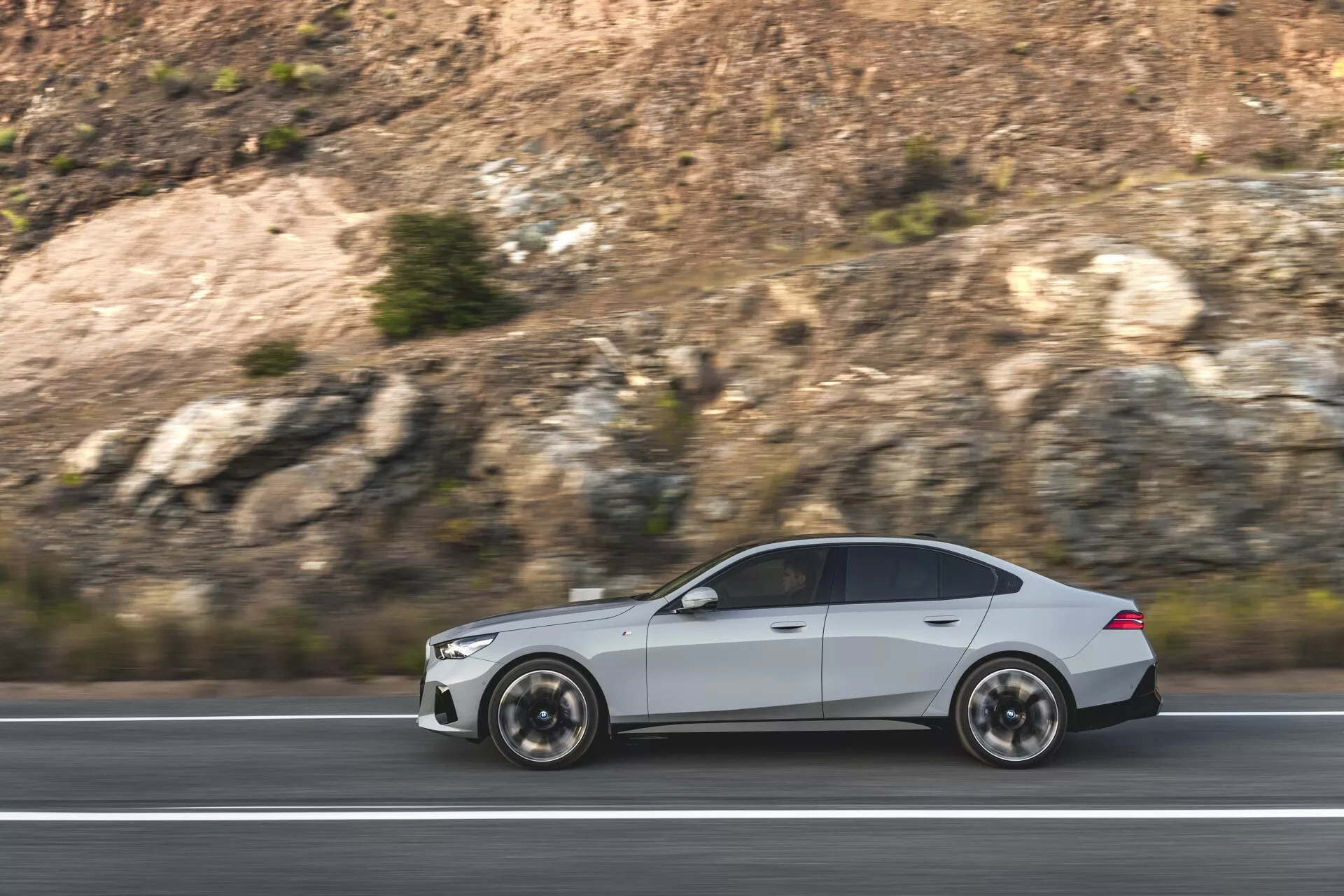 BMW Serie 5 (G60-G61) del 2023 in movimento, che mostra il suo design aerodinamico e l'estetica sportiva con ruote a più razze visibili e contorni del corpo eleganti, catturati dal lato su un'ambientazione autostradale