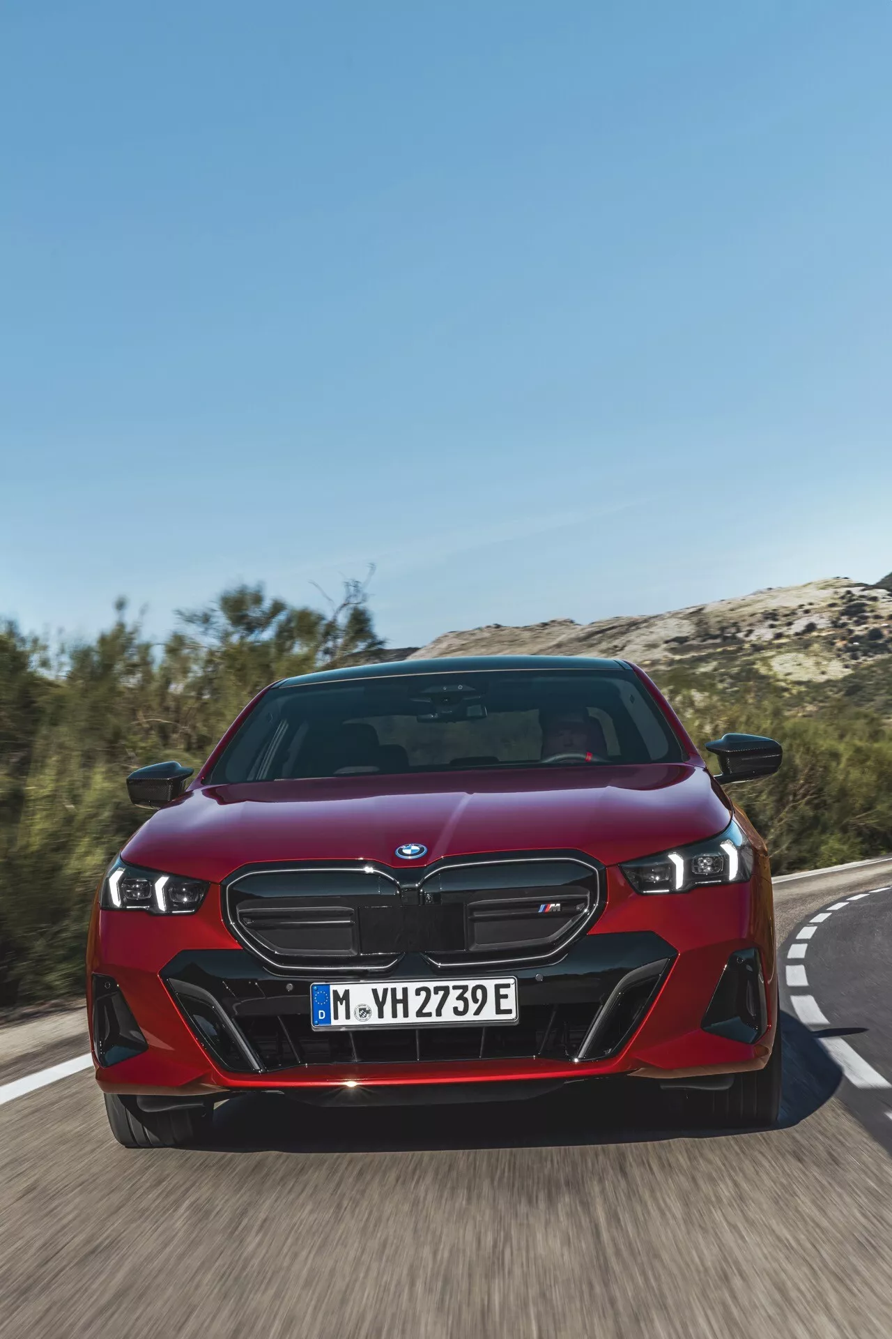 BMW Serie 5 (G60-G61) 2023 in rosso, che mostra la sua distintiva griglia a due reni, fari LED e fascia anteriore dinamica da un angolo basso frontale su una strada, evidenziando la velocità del veicolo e le avanzate funzionalità elettrificate iPerformance