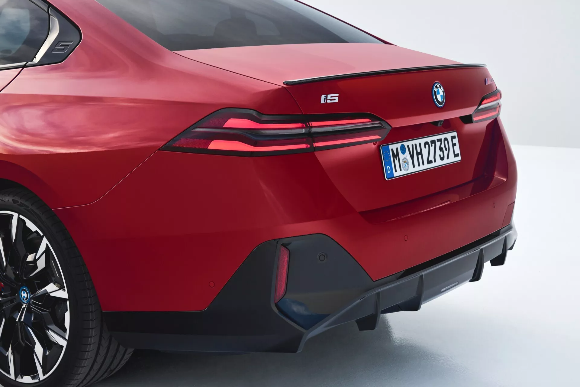 Vista angolare posteriore di una BMW i5 2023 in rosso che mostra il design sportivo della berlina, spoiler integrato, distintivo BMW prominente, fanali posteriori LED allungati che si estendono nei parafanghi posteriori, designazione del modello elettrico 'i5', paraurti posteriore aggressivo con diffusore decorativo e uscite di scarico false, in un profilo posteriore e laterale di tre quarti.