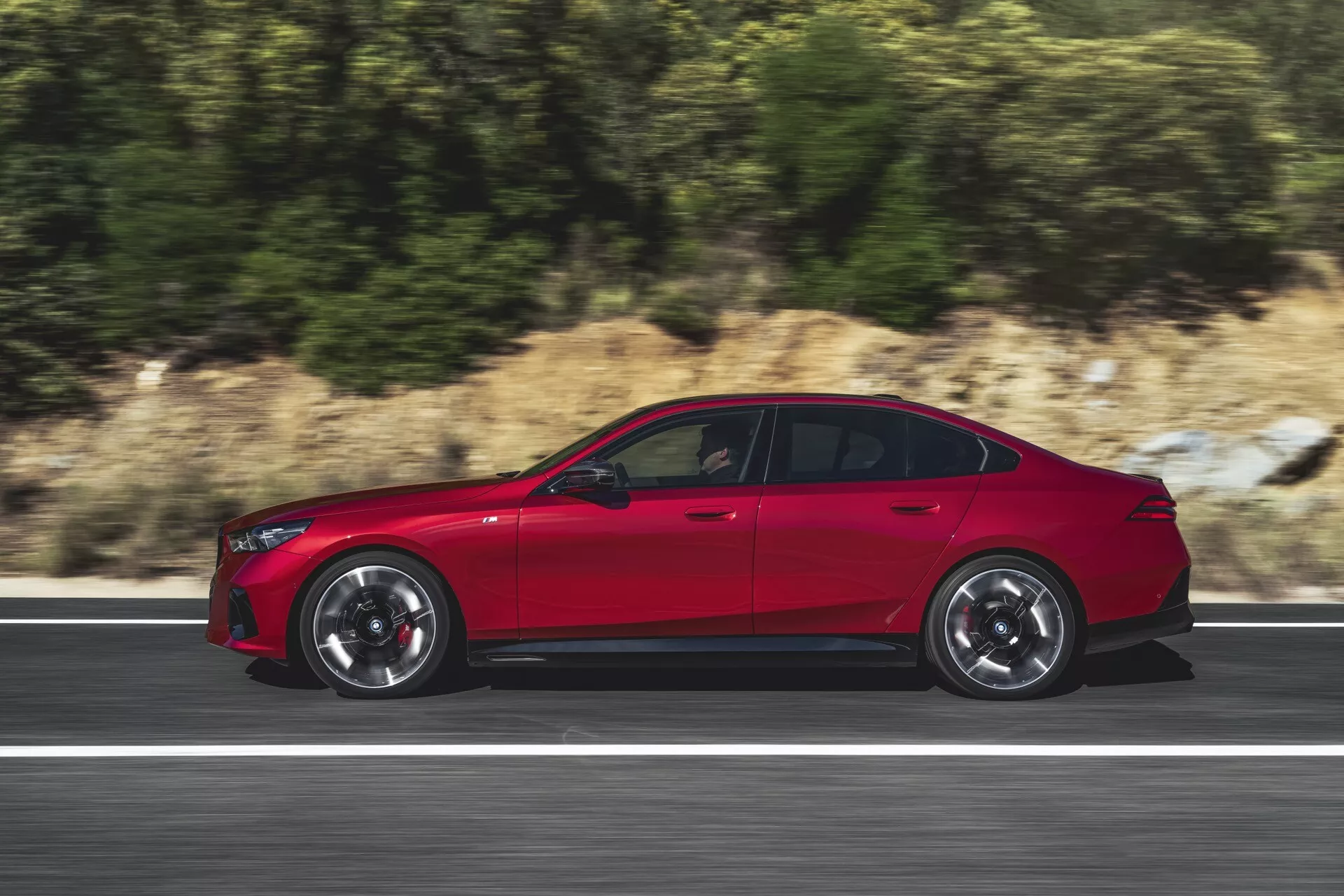 Profilo laterale dinamico della BMW Serie 5 (G60-G61) 2023 in movimento su strada che mostra design sportivo, grandi cerchi in lega a più razze e vernice rossa lucida che enfatizza l'aerodinamica e le caratteristiche tecnologiche avanzate