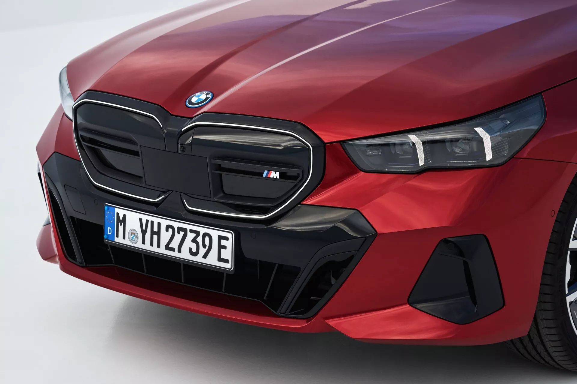 Vista angolata ravvicinata del lato anteriore sinistro di una BMW Serie 5 (G60-G61) del 2023 in rosso, caratterizzata dalla griglia renale iconica contrassegnata come modello 'M', gruppo ottico LED e elementi aerodinamici specializzati che suggeriscono capacità elettriche o ibride