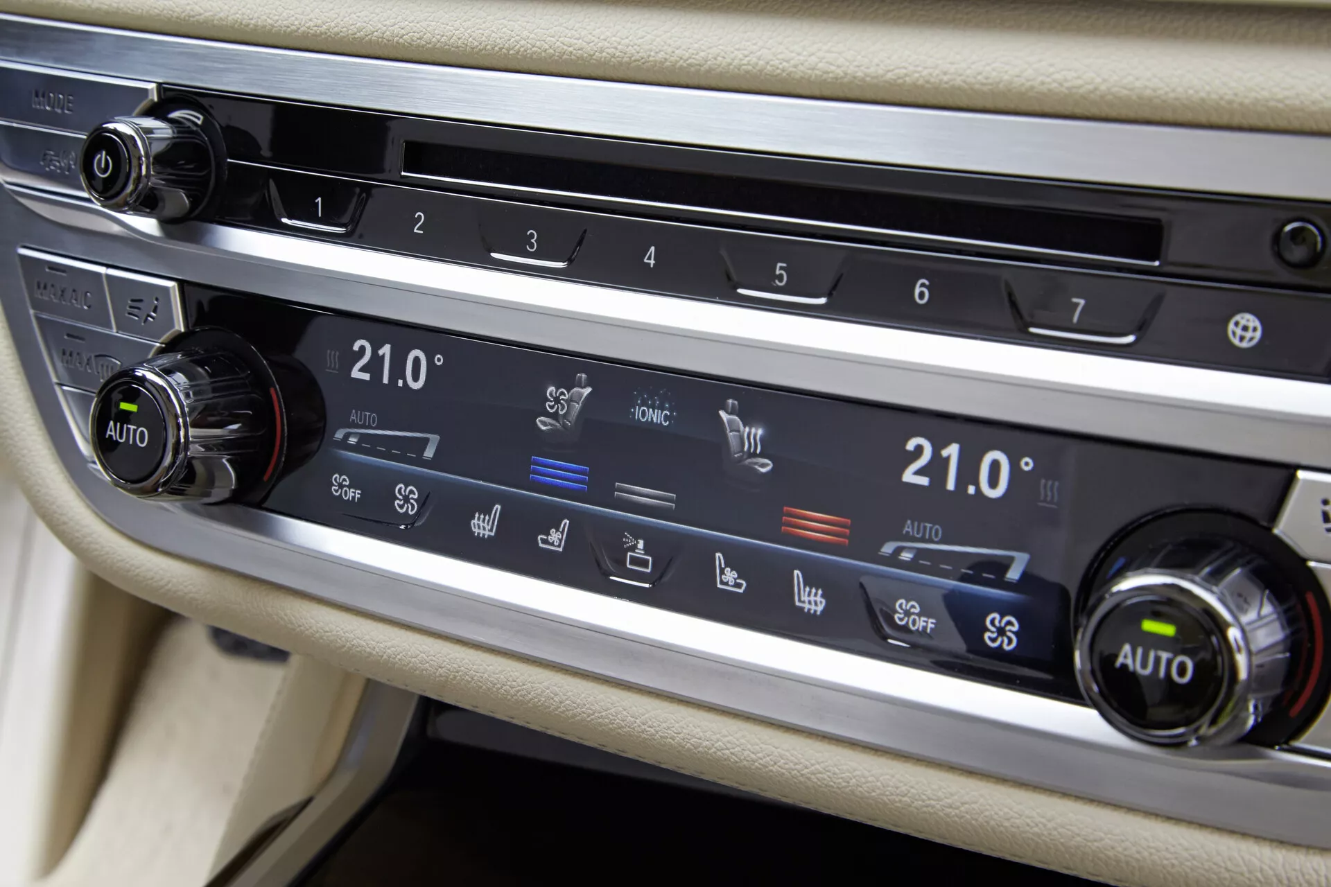 Vista ravvicinata del sistema di controllo del clima del BMW 7er-Reihe 2015 (G11-G12) sulla console centrale tra i sedili del conducente e del passeggero. Si concentra sul pannello orizzontale con due manopole e pulsanti per varie funzioni. Entrambe le manopole, impostate a 21,0°C e indicate come 'AUTO', suggeriscono un controllo automatico della temperatura a doppia zona. I pulsanti includono lo sbrinamento del lunotto posteriore, la circolazione dell'aria, lo sbrinamento del parabrezza e l'attivazione delle bocchette. Il sistema è incorniciato in una finitura metallica spazzolata contro un interno di colore chiaro, con una possibile striscia di display o superficie tattile sopra.