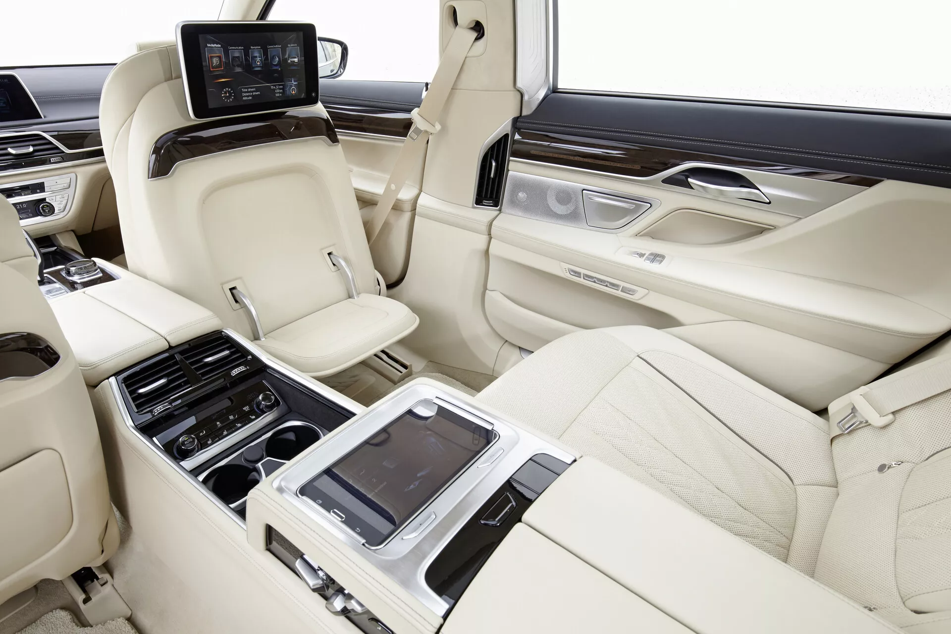 Immagine dell'interno della cabina posteriore della BMW 7er-reihe (G11-G12) del 2015, che cattura l'area dei sedili spaziosa e lussuosa da una prospettiva destra. I sedili in pelle di colore chiaro in configurazione 2+1, con design sagomato per comfort e supporto, poggiatesta e capacità di reclinazione individuale sono visibili. Una console centrale con vari comandi e possibilmente un'interfaccia touch screen per le impostazioni dei sedili, il controllo del clima e i multimedia è in evidenza. I pannelli delle porte includono rifiniture in legno e accenti metallici spazzolati con comandi aggiuntivi. Monitor individuali sono montati sul retro dei sedili anteriori per l'intrattenimento e un pannello di controllo posteriore con uno schermo è posizionato dietro la console centrale. Gli interni del veicolo mostrano avanzate regolazioni dei sedili, controllo del clima e sistemi di intrattenimento all'interno di finiture di alta gamma in pelle, legno e metallo.