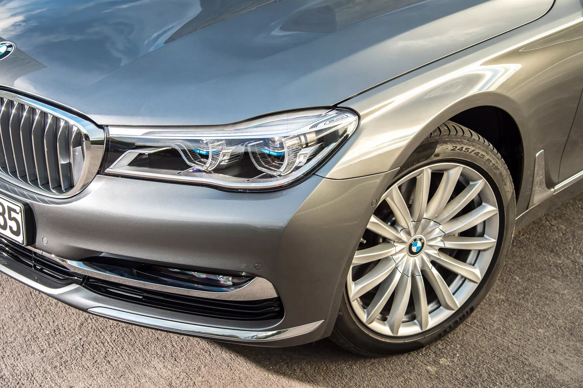 Inquadratura ravvicinata angolata di una berlina di lusso argento BMW 7er-reihe 2015 (G11-G12), con particolare attenzione alla sezione anteriore sinistra. Mette in evidenza il paraurti anteriore, il faro sinistro, il cofano, il parafango e il cerchio in lega a più razze. Fari LED complessi con luci di marcia diurna distintive e piccoli quadrati blu indicano sistemi di illuminazione avanzati. La griglia anteriore parzialmente visibile con lamelle verticali cromate e una telecamera o un sensore nelle vicinanze suggeriscono tecnologia di assistenza alla guida. Le prese d'aria sul paraurti anteriore e la finitura della vernice metallica sottolineano ulteriormente il design lussuoso e aerodinamico dell'auto.