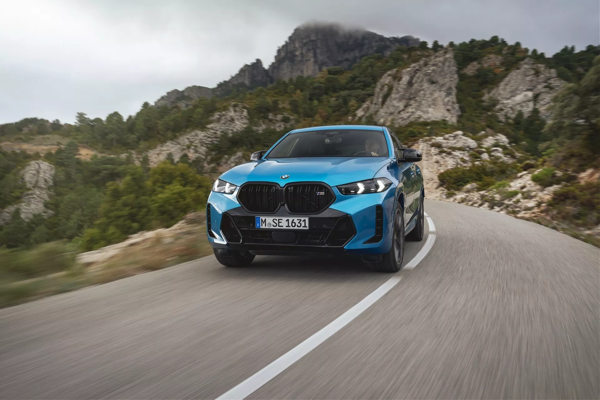 BMW X6 2023 (G06-F96-facelift) in blu vibrante, che mostra prestazioni di lusso moderne su una strada di montagna tortuosa con sfocatura dinamica, fari LED avanzati, elementi di design elegante e ruote sportive, su uno sfondo di montagne aspre.