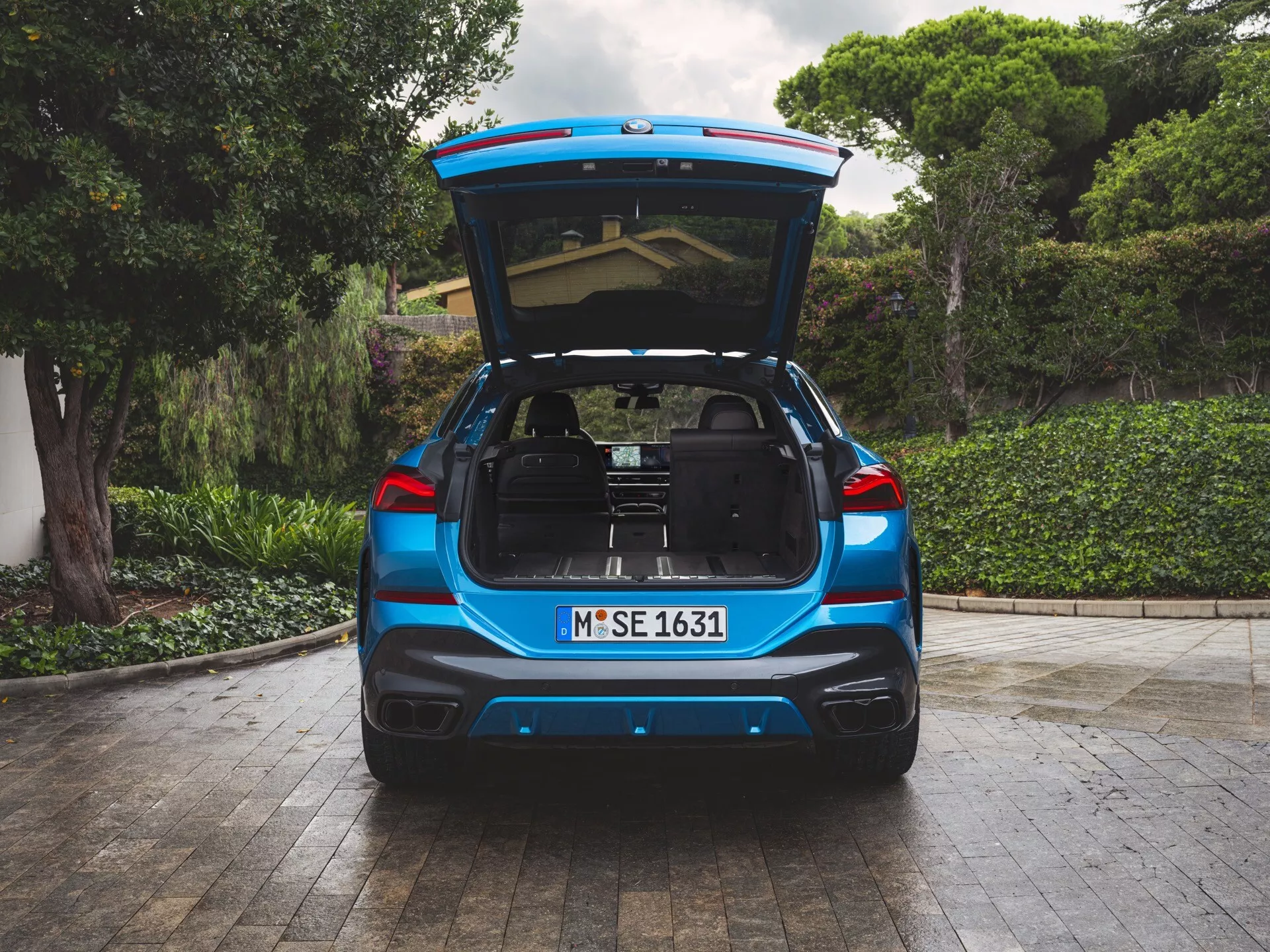 Vista posteriore del BMW X6 2023 (G06-F96-Facelift) con vernice metalizzata blu, bagagliaio aperto che rivela un'ampia area di carico, parcheggiato su pavimentazione in pietra con vegetazione e parte di una casa sullo sfondo, dotato di fanali posteriori LED, doppi scarichi e possibili tecnologie assistenza parcheggio