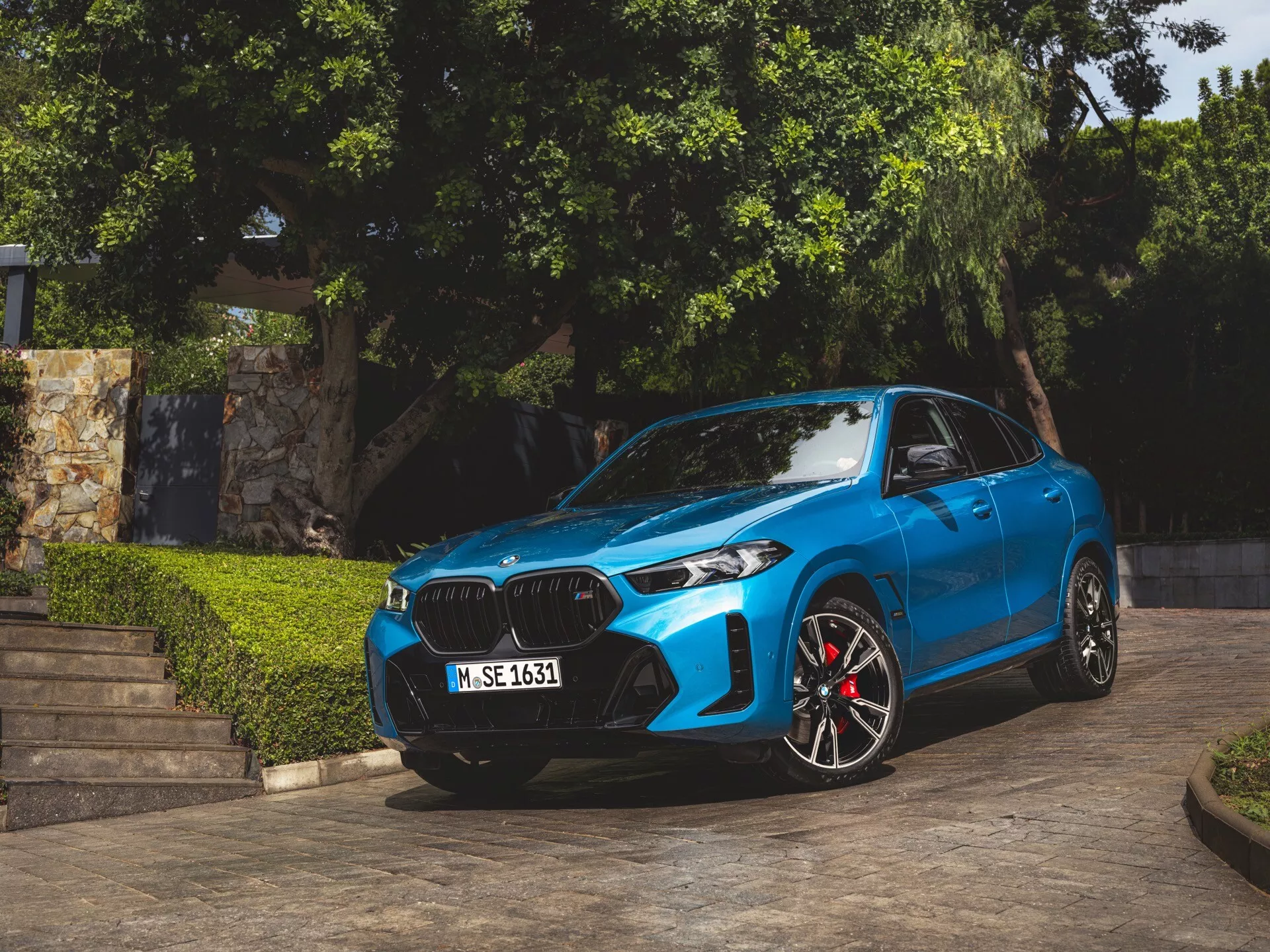 BMW X6 2023 (G06-F96-Facelift) di colore blu, parcheggiata ad angolo mostrando la sua facciata anteriore, griglia a forma di rene, fari LED o laser, ruote sportive in lega con pinze dei freni rosse, linee contornate sul cofano e specchietti laterali aerodinamici con indicatori di direzione integrati, catturata alla luce naturale del giorno con vegetazione e gradini architettonici sullo sfondo