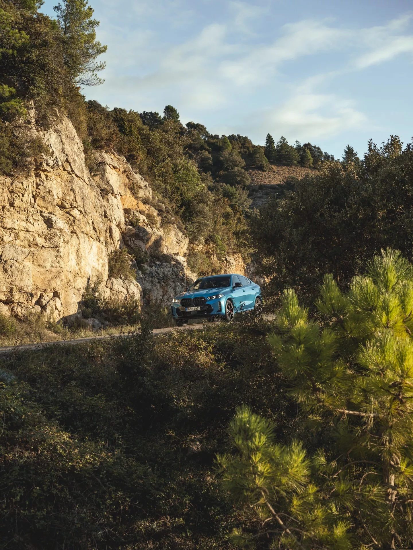 BMW X6 2023 (restyling G06-F96) in vista frontale a tre quarti su strada montana tortuosa circondata da bosco, dimostrando capacità fuoristrada e design moderno
