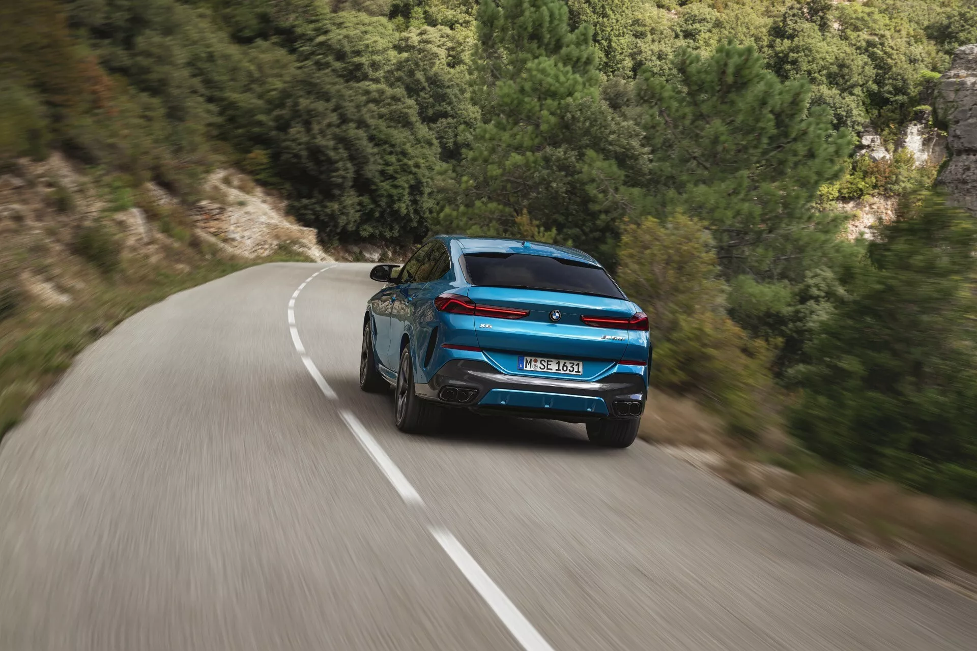 Vista posteriore dinamica di una BMW X6 2023 (G06-F96-Facelift) blu, con design sportivo e fanali posteriori moderni su strada tortuosa in mezzo a un paesaggio naturale