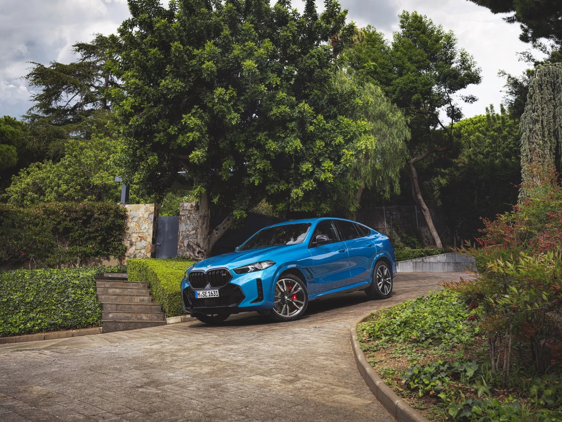 BMW X6 2023 (G06-F96-Restyling) in blu, vista frontale tre quarti, parcheggiata su un vialetto con verde circostante, mostrando paraurti anteriore, griglia, cofano, parabrezza, profilo laterale del conducente, fari LED, ruote sportive con pinze freno rosse e design aerodinamico