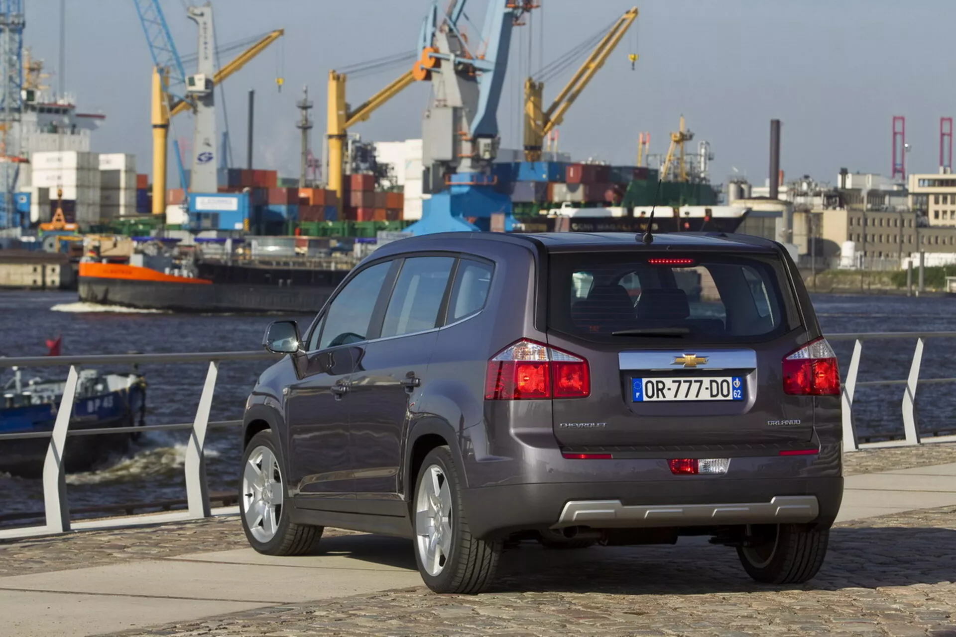 Vista posteriore a tre quarti di una Chevrolet Orlando 2011 in un molo sul lungomare, che evidenzia la sua carrozzeria SUV compatta, configurazione a cinque porte, sistema di scarico doppio, sensori di assistenza al parcheggio sul paraurti e ruote in lega a più razze