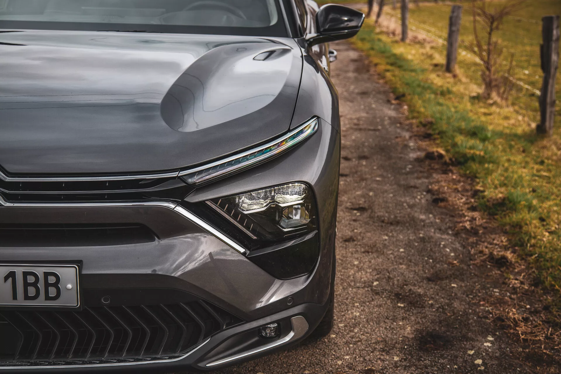 Primo piano del Citroen C5-X 2022 che mette in mostra il design elegante del lato anteriore sinistro, incluse la cappuccia sagomata, la griglia pronunciata con linee orizzontali, il sistema di fari LED avanzato e il paraurti anteriore aerodinamico con prese d'aria, su sfondo naturale sfocato a bordo strada.