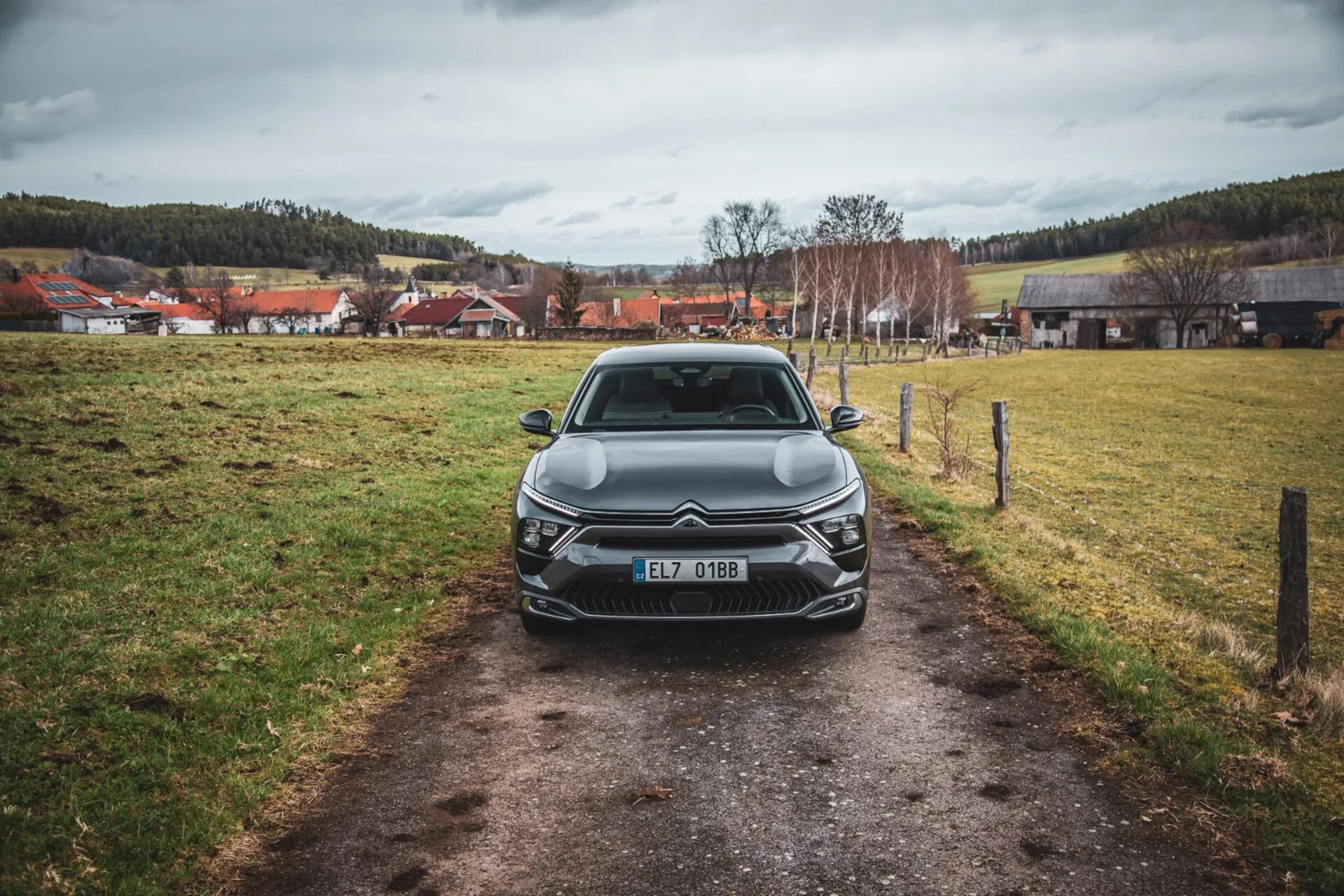 Vista frontale di una Citroën C5-X 2022 su un sentiero stretto in un campo erboso sotto un cielo coperto, che mette in mostra il suo design elegante, griglia intricata con emblema, unità fari LED angolari, linee scolpite sul cofano e elementi di illuminazione anteriore aggiuntivi, registrato nella Repubblica Ceca.