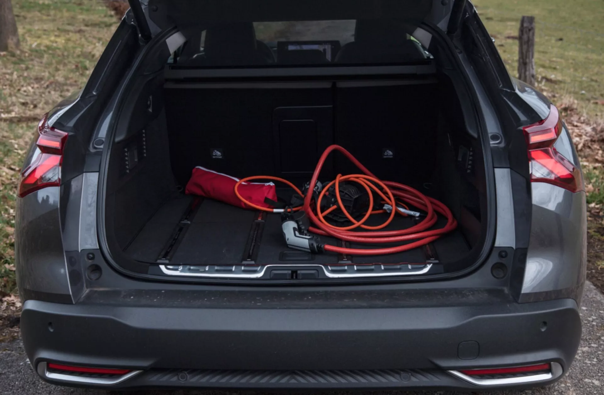 Vista dello spazio bagagliaio posteriore della Citroen C5-X 2022 con vano aperto che mostra area di carico spaziosa, borsa rossa e nera, cavo di prolunga arancione, recessi per stoccaggio aggiuntivo, profilo protettivo, e sensori di parcheggio, in luce naturale all'aperto
