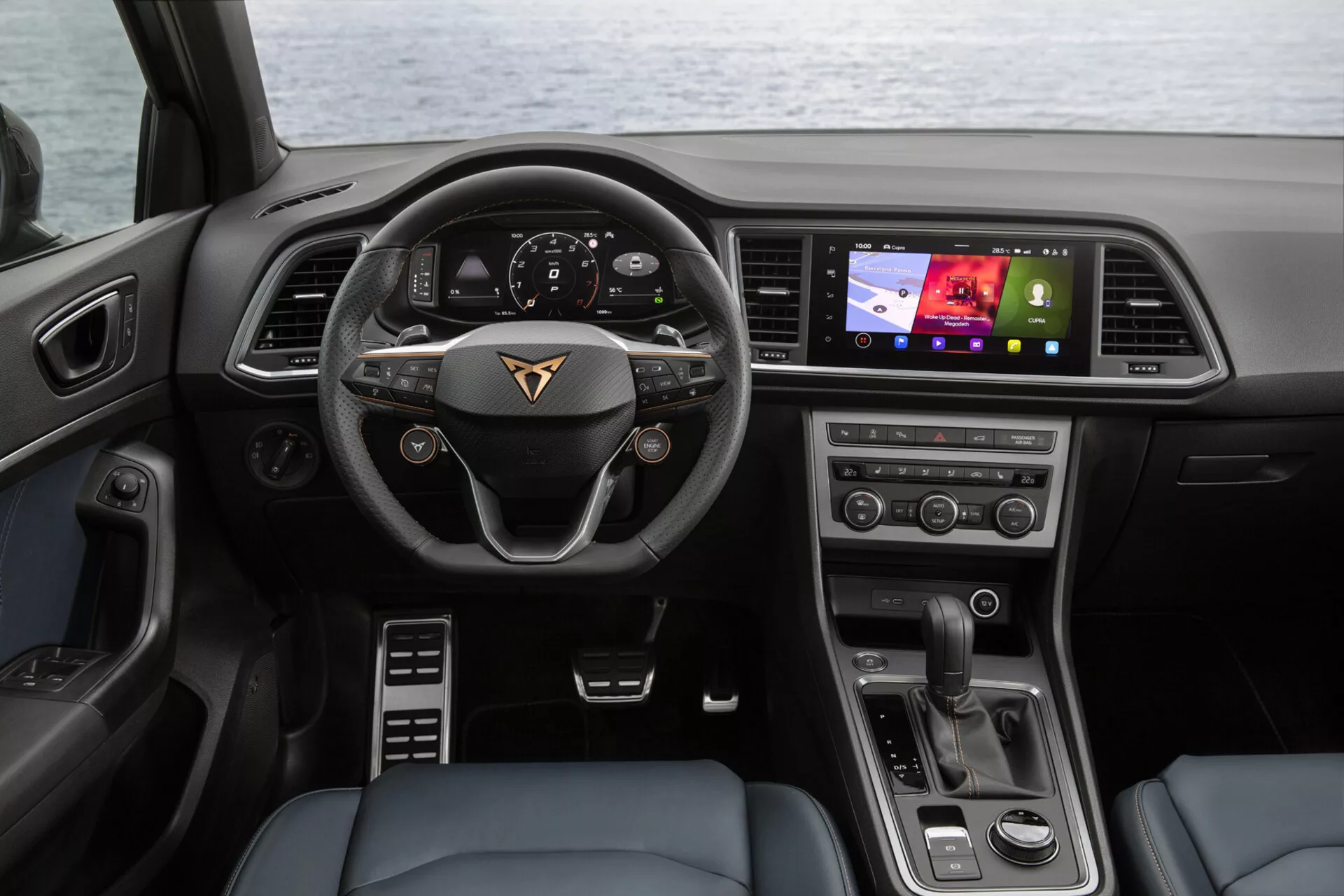 Vista interna della Cupra Ateca 2020 dal sedile posteriore che mostra la plancia, il volante multifunzionale con emblema del produttore, il cruscotto digitale, il touchscreen dell'infotainment orientato verso il guidatore, i pulsanti per il controllo del clima e il selettore di marcia moderno