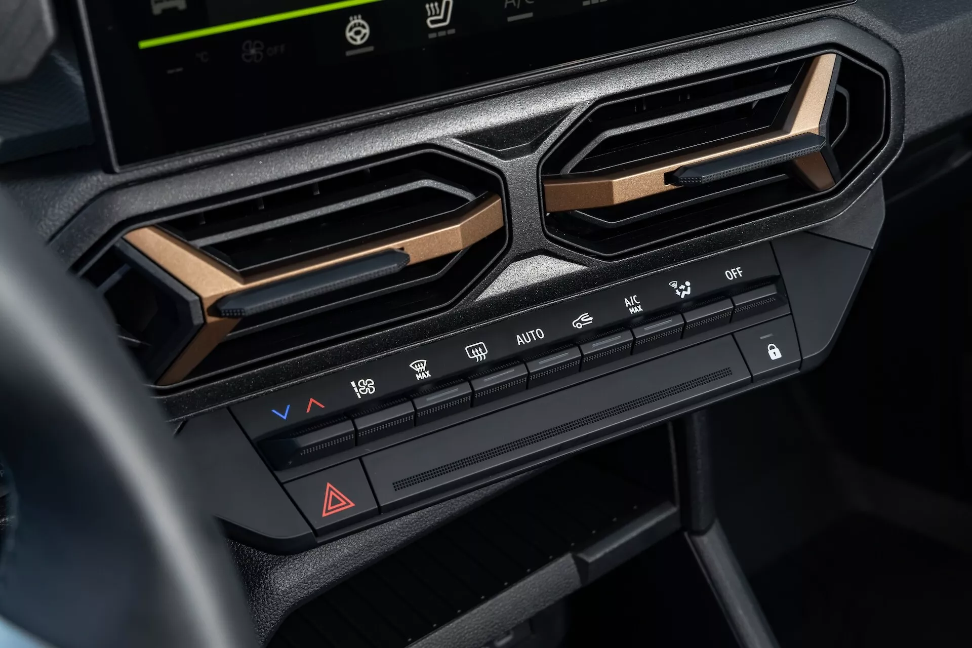 Vista ravvicinata del sistema di controllo clima su Dacia Duster 3ª generazione 2024 nel cruscotto centrale sotto lo schermo dell'infotainment, evidenziando regolazioni di temperatura e velocità della ventola, funzioni di distribuzione dell'aria, caratteristiche di sbrinamento, pulsante delle luci di emergenza e opzioni di aria condizionata, incorniciate da bocchette d'aria angolari e accentate con rifiniture metalliche per un'estetica interna moderna