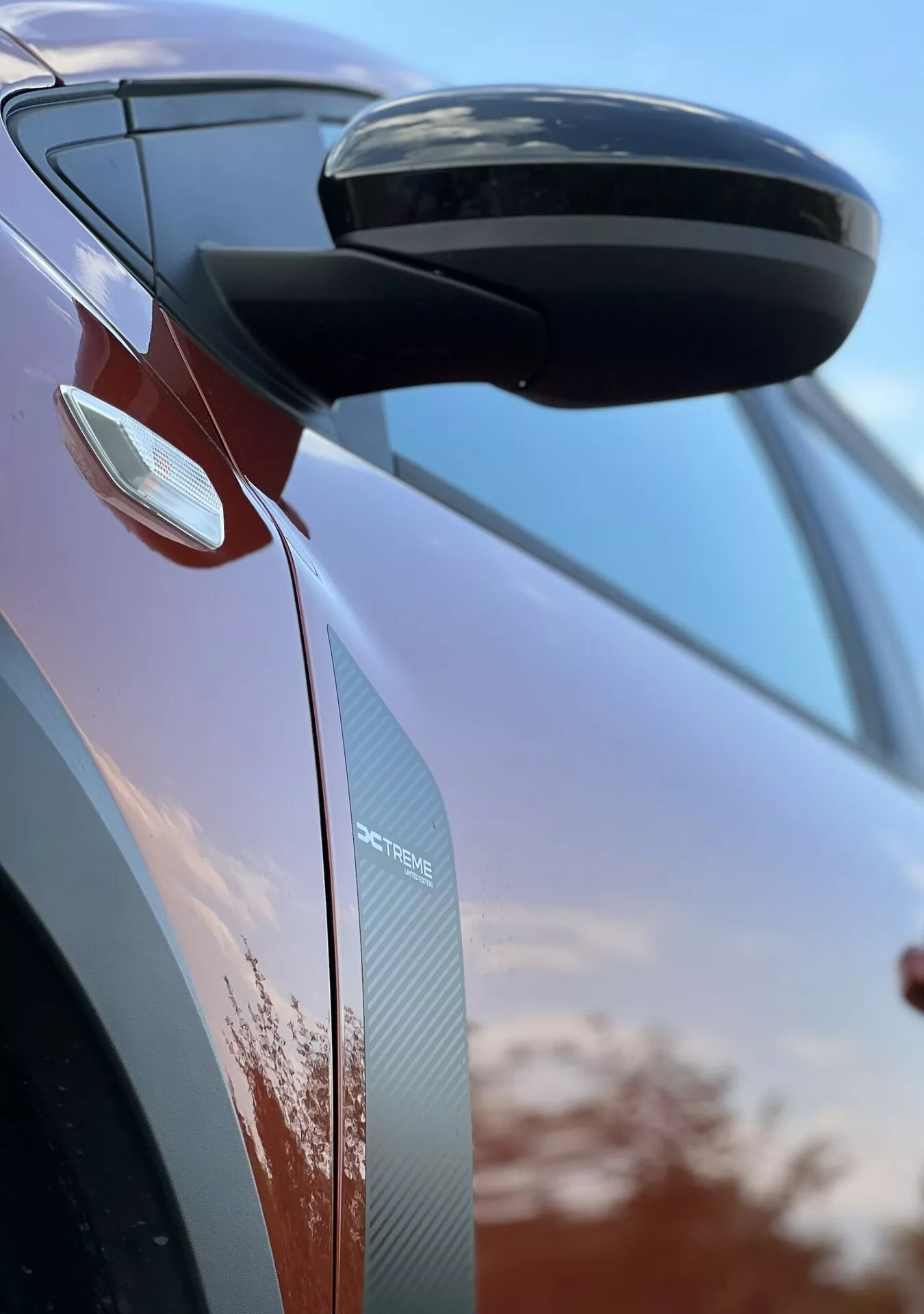 Dettaglio ravvicinato del Dacia Jogger 2022 che si concentra sullo specchietto retrovisore laterale e sul design della porta, mettendo in mostra caratteristiche aerodinamiche, indicatori di direzione integrati e texture in fibra di carbonio su uno sfondo di cielo