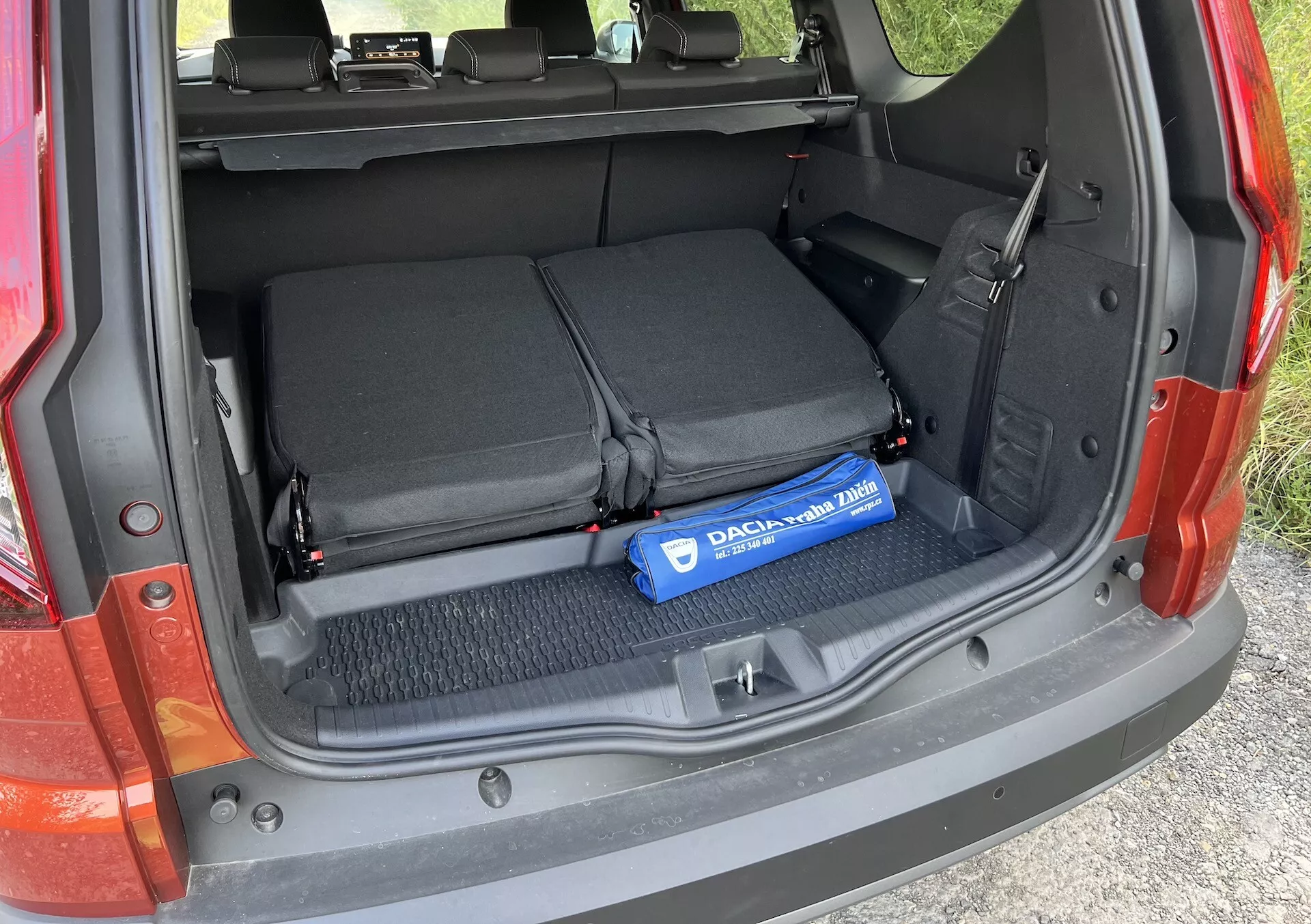 Dettaglio del bagagliaio della Dacia Jogger 2022 che mostra l'area di carico aperta con i sedili posteriori abbattuti, cinghie tessili per fissare oggetti e un cilindro blu Decaux sul pavimento rivestito in moquette