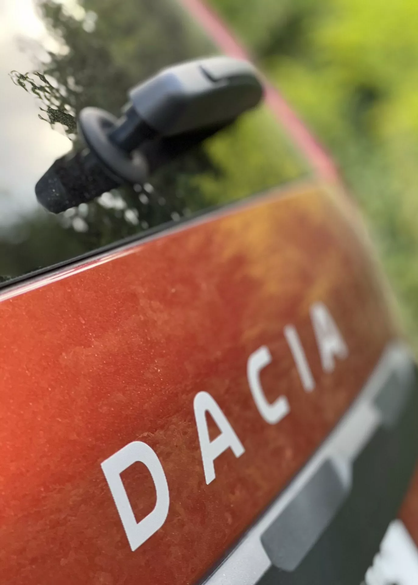 Vista ravvicinata del pannello posteriore della Dacia Jogger 2022 con il nome DACIA in lettere maiuscole bianche su una verniciatura metallica arancione-rossa con macchie e sporco, suggerendo l'uso all'aperto e la durata, situata in un ambiente naturale con uno specchietto laterale sfocato che indica caratteristiche moderne dello specchio.