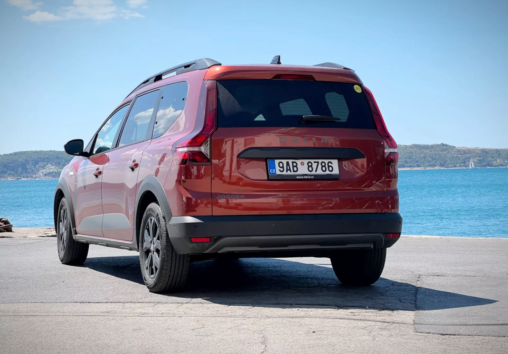 Dacia Jogger 2022 in rosso, ripresa da un angolo posteriore di tre quarti che mostra il lato e il design del portellone dell'SUV, parcheggiata vicino a un corpo d'acqua con orizzonte sullo sfondo, evidenziando le sue ruote in lega a più razze, fanali posteriori LED e spoiler posteriore aerodinamico