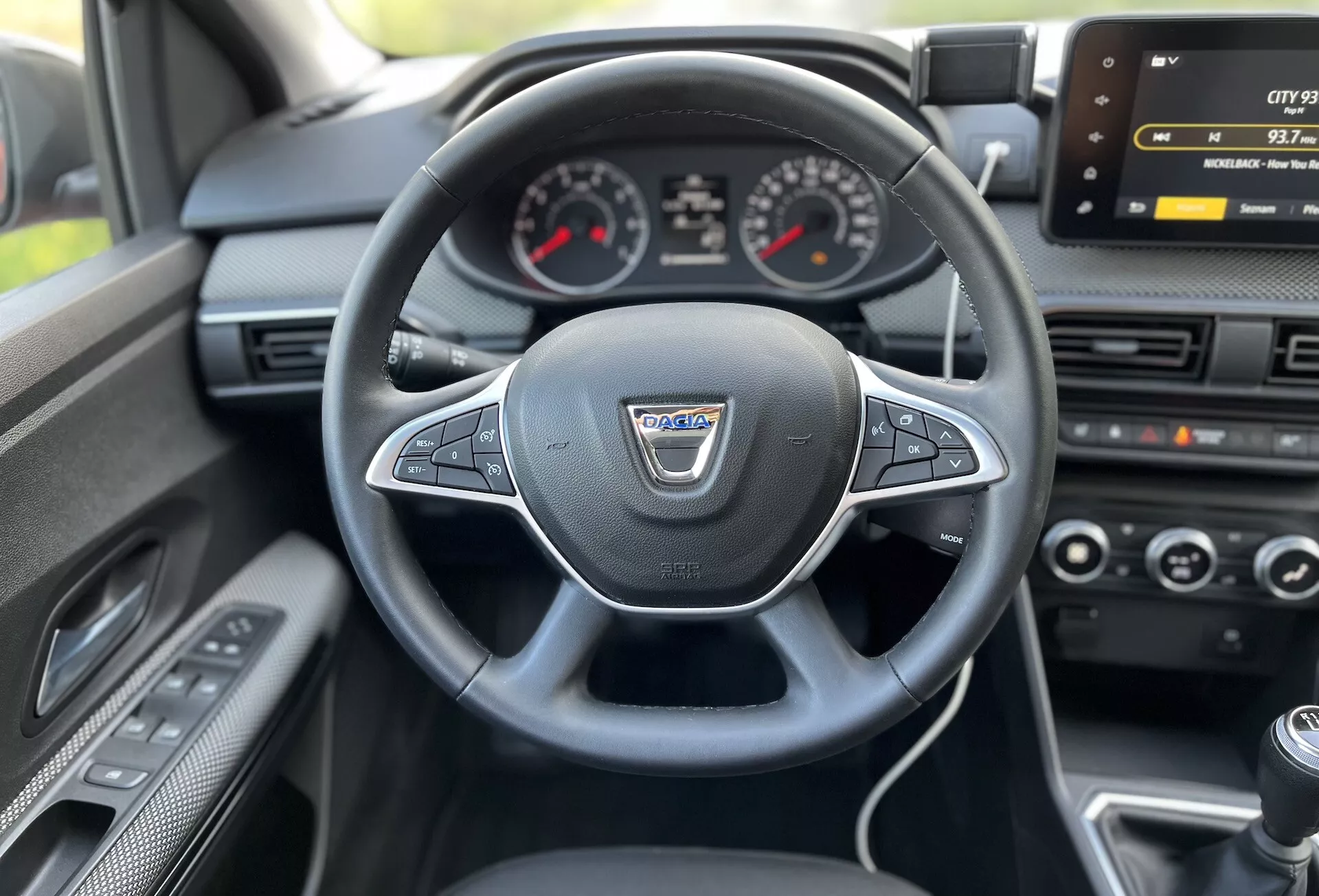 Vista interna di una Dacia Jogger 2022 dal sedile del conducente che mostra il volante multifunzione rivestito in pelle, cruscotto analogico con display digitale centrale, schermo infotainment, controllo del clima e leva del cambio manuale